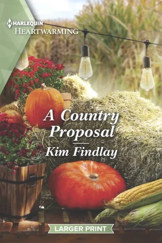 Cover art for author Kim Findlay’s romance novel “A Country Proposal.” A Harvest festival table is arranged in the foreground.  There is a hay bale a string of old-fashioned light bulbs for decoration.  Hay fields are blurry in the background