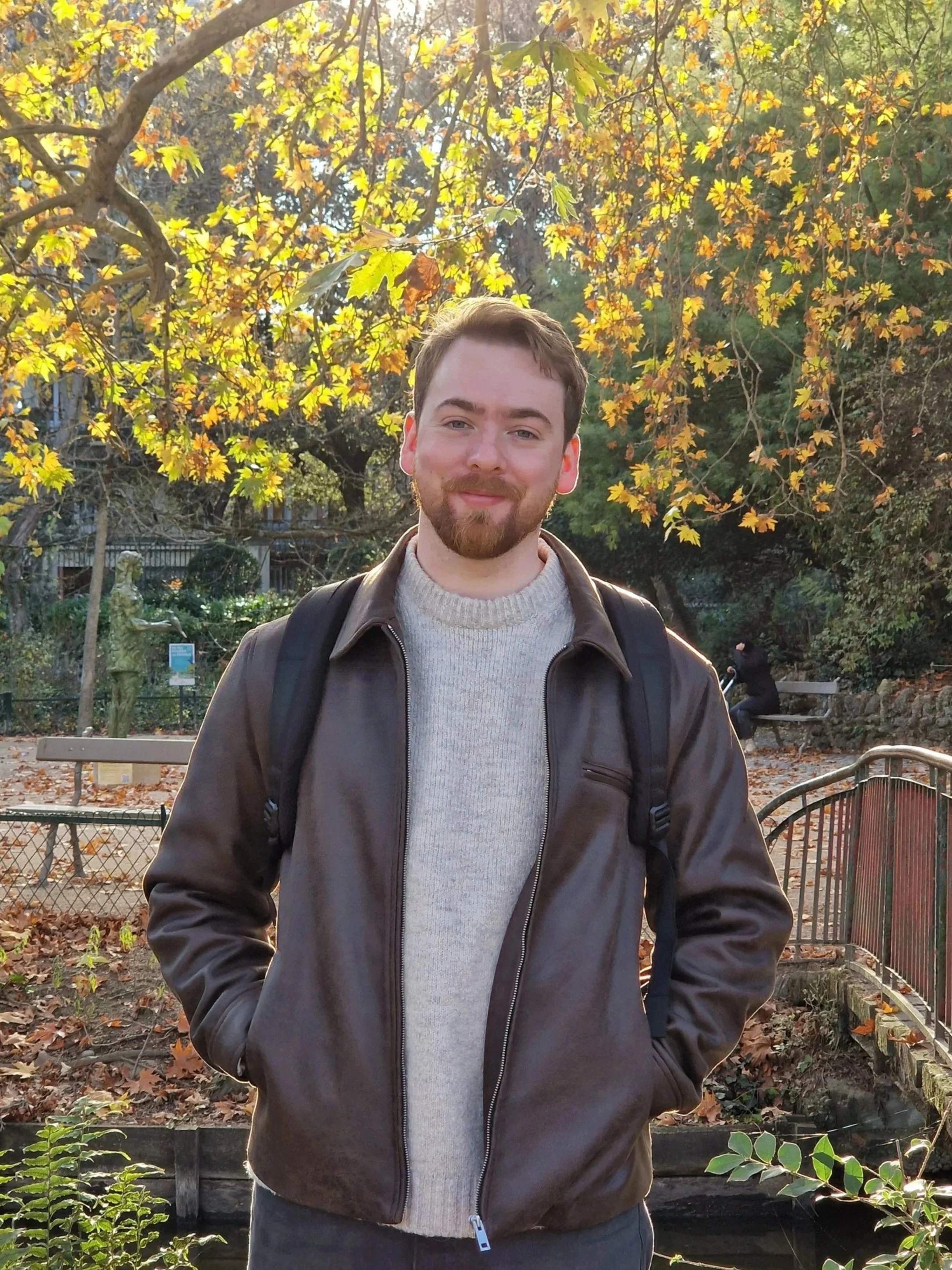 A young man with a beard and short brown hair standing outdoors during autumn, surrounded by yellow and orange leaves, wearing a brown leather jacket over a cream knit sweater, with a black backpack.