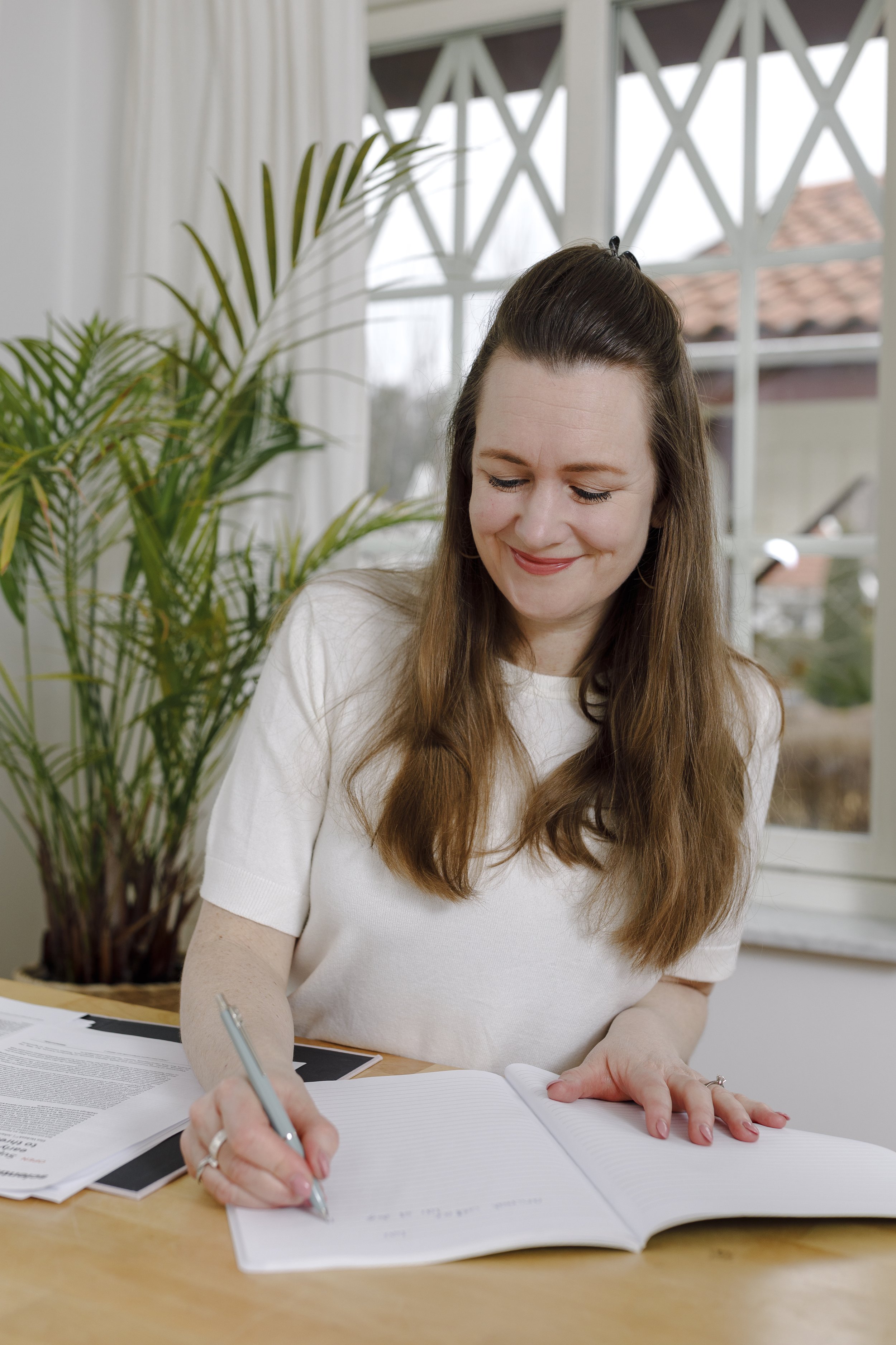 A woman with long brown hair sitting at a wooden desk, smiling, as she writes in a notebook with a silver pen. There are papers on the desk and a green potted plant behind her, with a window in the background.