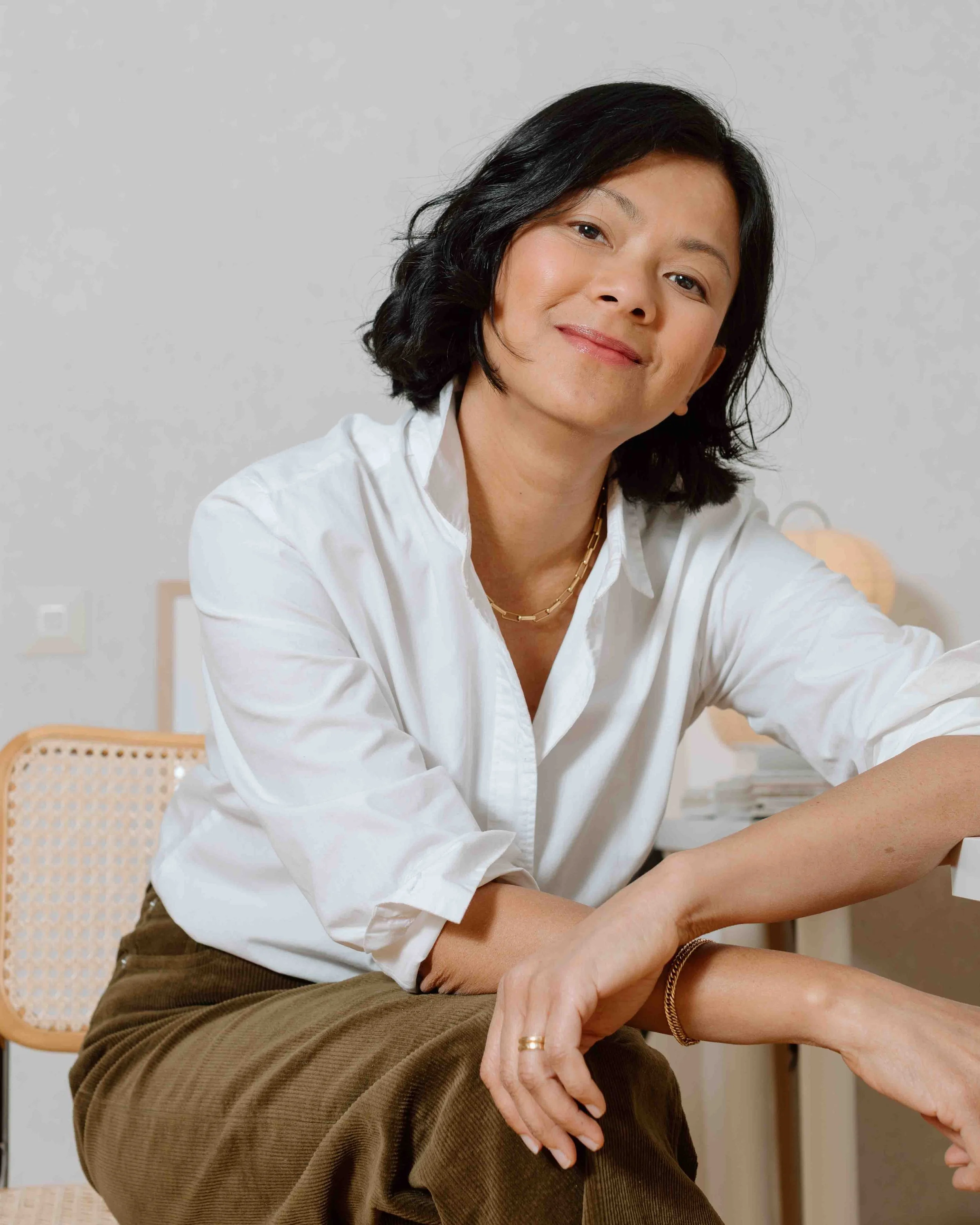 A woman with black hair in a white shirt and brown pants sitting at a desk in a bright room, smiling at the camera.