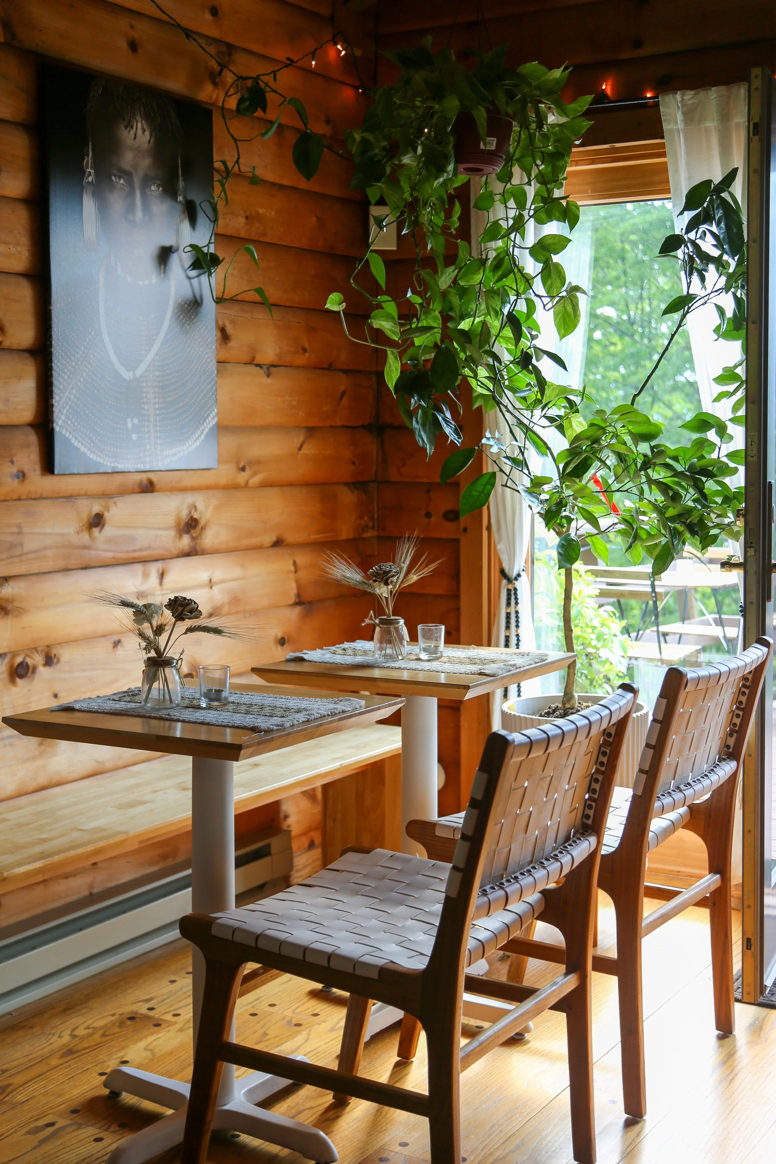 Cozy wooden dining area with two modern chairs and small tables adorned with table mats, dried flowers, and candles. A potted plant hangs near a window, and a black-and-white portrait is displayed on the wooden wall.