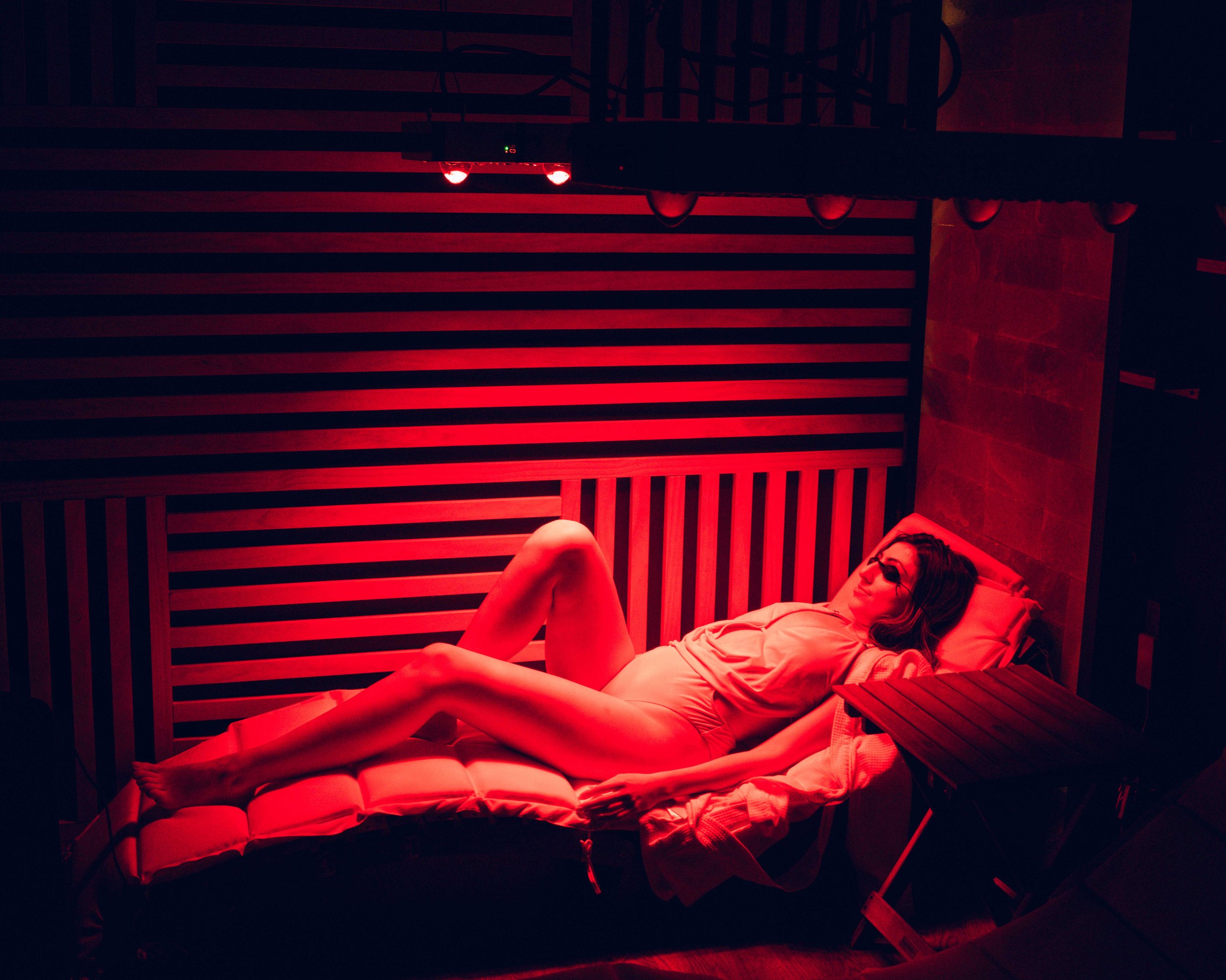 A wooden sauna cabin with glass door on a deck surrounded by trees, winter scene with a light dusting of snow and a black Sovilo branded tub nearby.