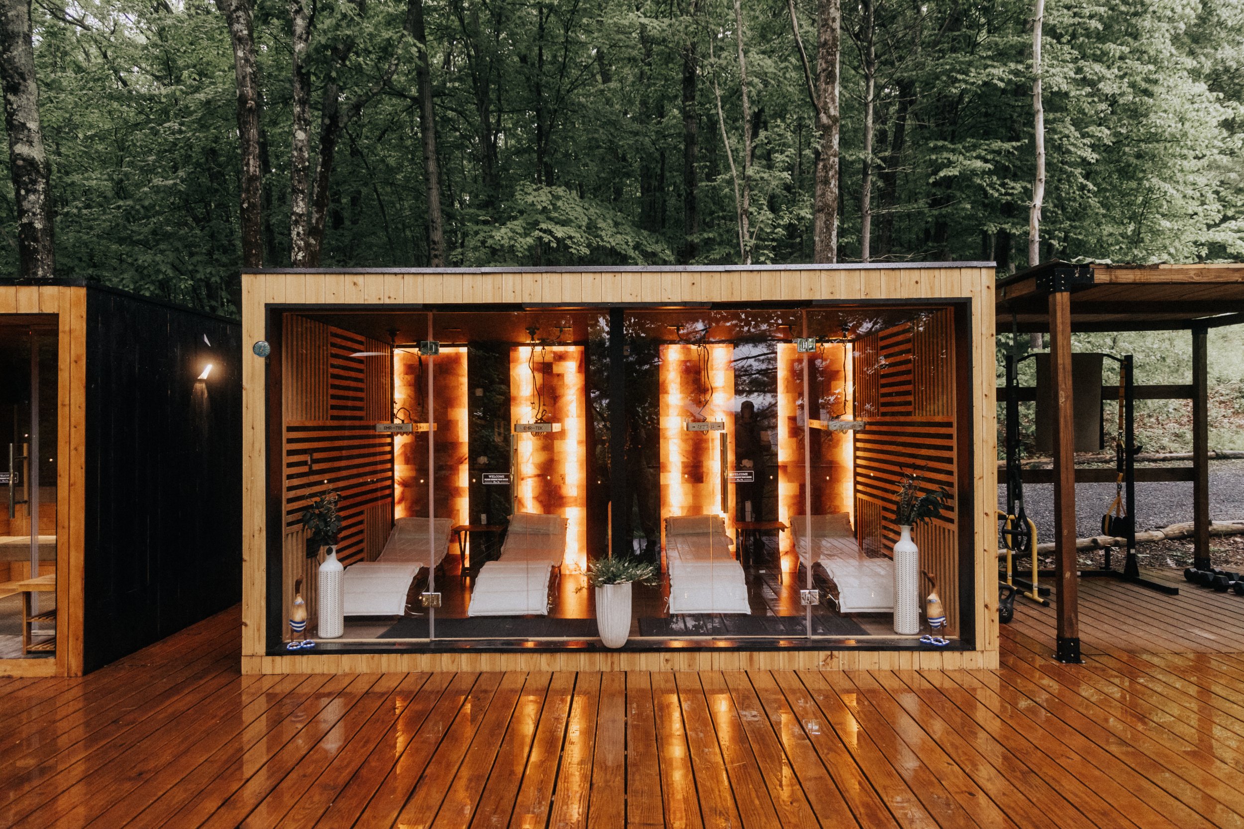 A wooden sauna cabin with glass door on a deck surrounded by trees, winter scene with a light dusting of snow and a black Sovilo branded tub nearby.