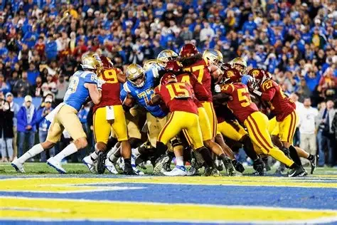 Line of football players from opposing teams during a game, with one team in blue and beige and the other in red and yellow spots, on the field with a crowd in the background.
