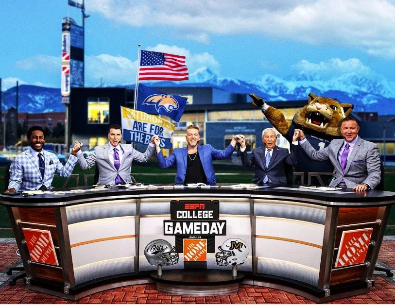 Five men, some in suits, standing behind a desk with ESPN College Gameday signage, holding a flag, and raising their fists in celebration, with a mountain background and large mascot head on the right.