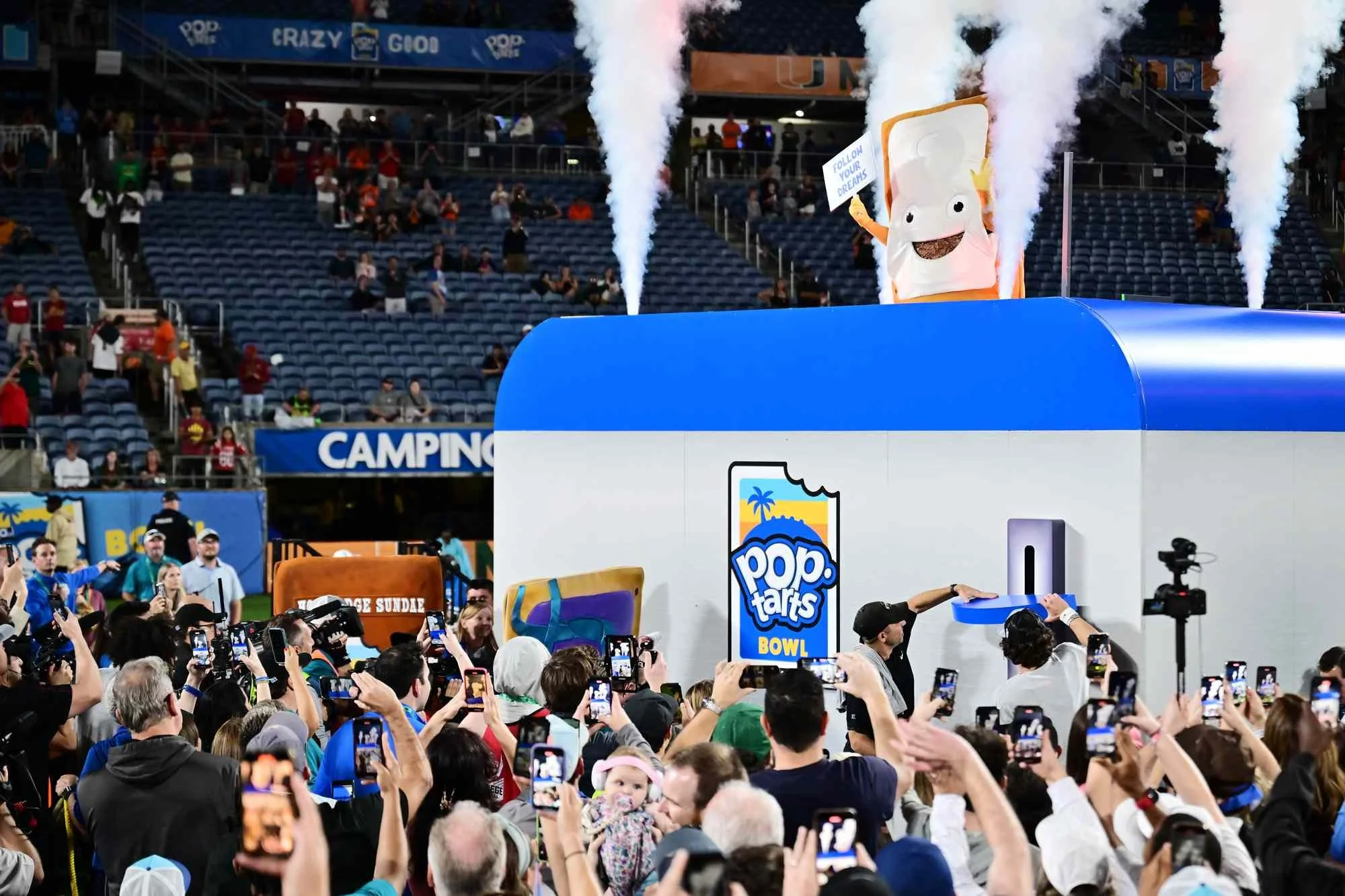 Crowd at a tattooed T-shirt display at a sports stadium, with a mascot in a popcorn bag costume and smoke effects in the background.