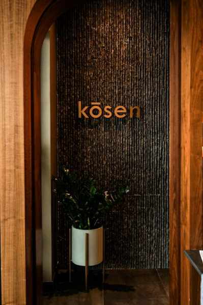 A view through a wooden archway of the Kōsen restaurant entrance, featuring a gold "kōsen" logo on a dark, textured stone wall with a green plant in a white planter.