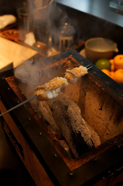 Two pieces of white fish on skewers being grilled over glowing Binchotan charcoal in a traditional Japanese hearth, with smoke rising in a dark kitchen.