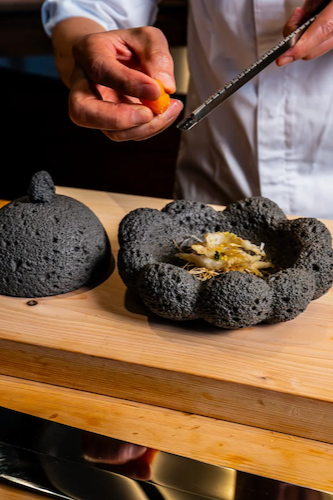 Close-up of a chef’s hands using a microplane to grate fresh citrus or egg yolk over a dish served in a unique, textured black volcanic stone bowl.