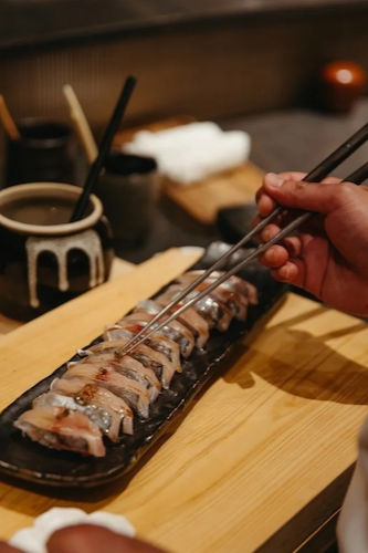 Close-up of a chef’s hands using long metal chopsticks to apply a final garnish to a row of fresh silver-skinned fish nigiri (likely Kohada or Aji) resting on a black ceramic platter over a light wood cutting board.