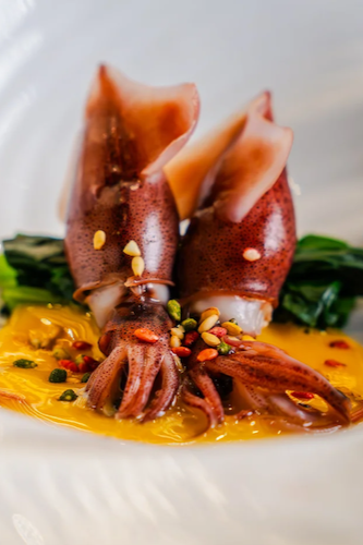 Two Firefly Squid (Hotaru Ika) served over bright green vegetables and a vibrant yellow miso-based sauce, garnished with small seeds and petals.