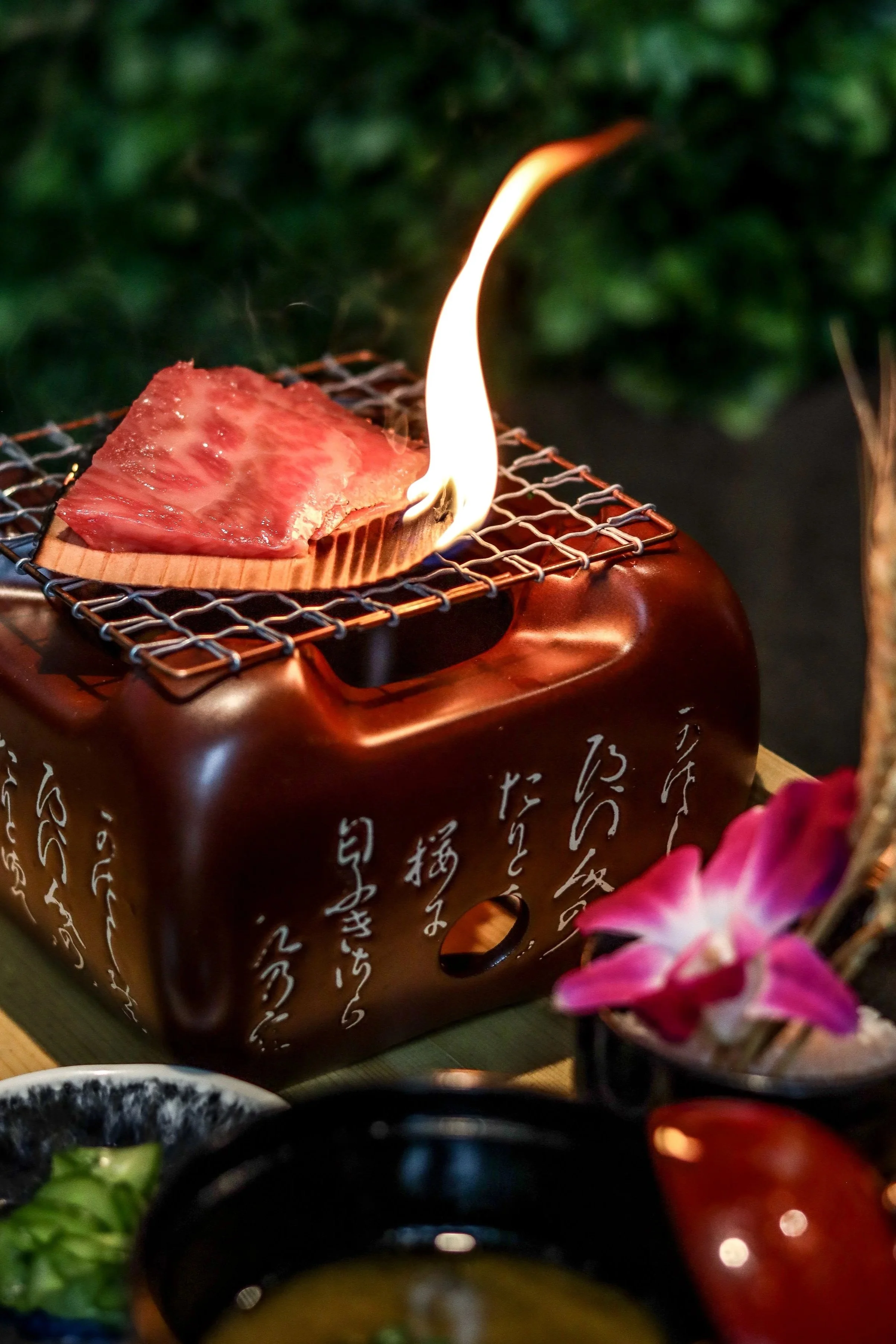 A premium slice of marbled A5 Wagyu beef sits on a piece of cedar wood atop a small, traditional Japanese ceramic grill (shichirin) against a lush green background at Kōsen in Tampa.