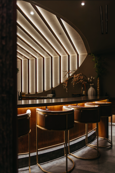 A wide shot of the elegant omakase bar at Kōsen in Tampa, featuring curved leather bar stools with gold frames. The background is dominated by the signature "Ray of Light" architectural wood ribbing and a minimalist floral arrangement.
