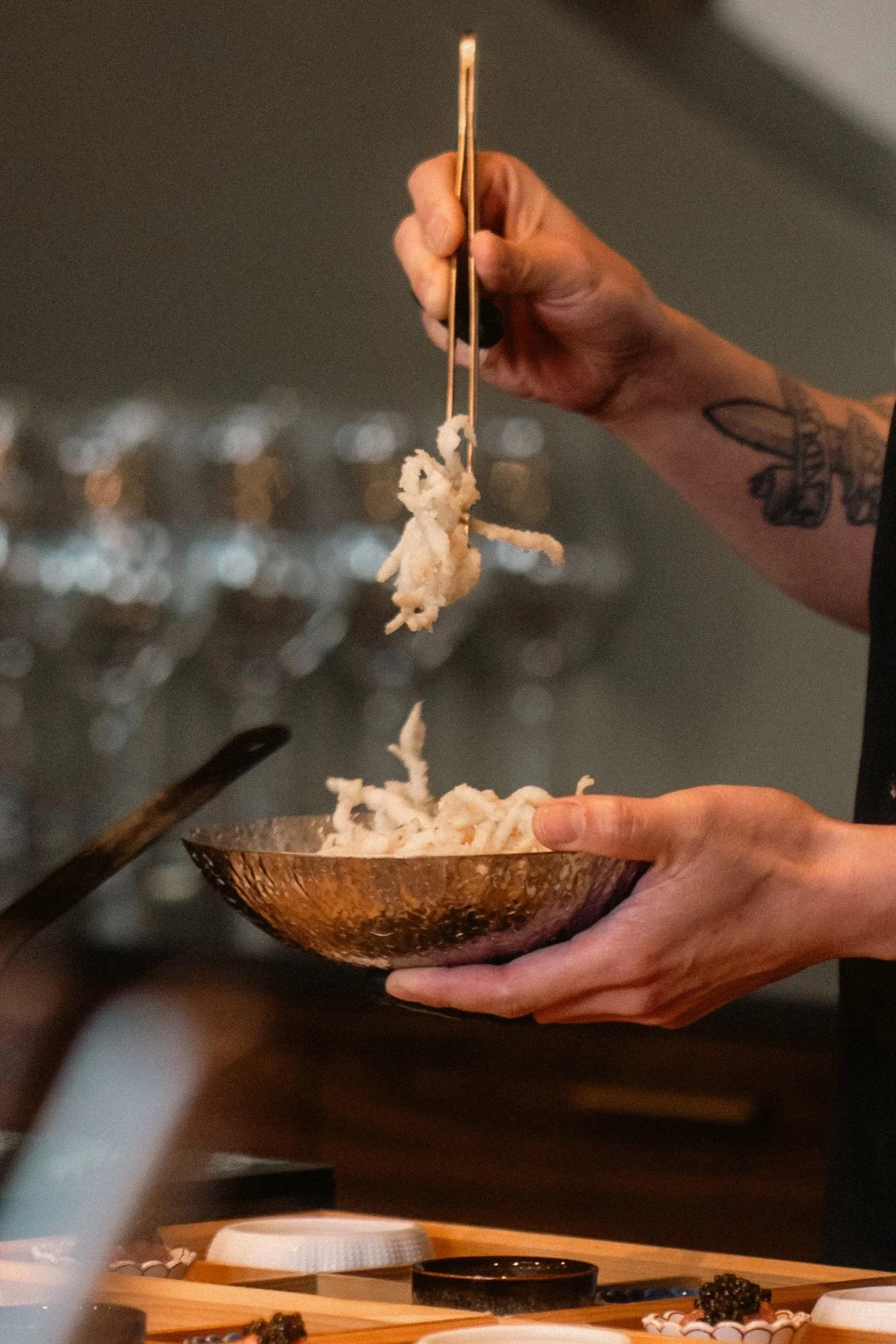 side view of hand holding a copper bowl of dried fish with the other hand picking up the dried fish with gold tongs
