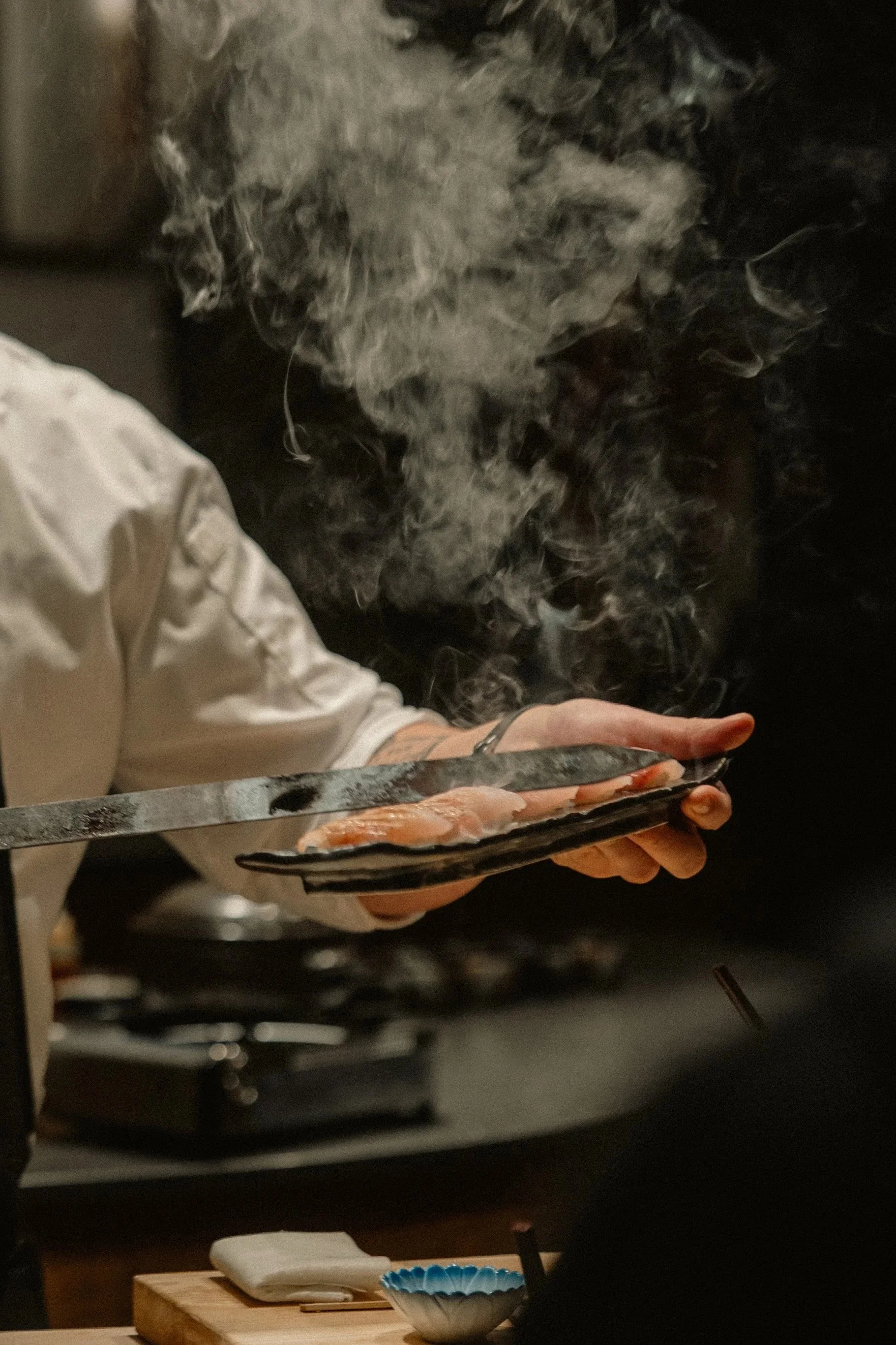 chef in whit coat searing a plate of nigiri with the blade of a hot sword, smoke arising
