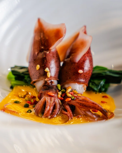 Two Firefly Squid (Hotaru Ika) served over bright green vegetables and a vibrant yellow miso-based sauce, garnished with small seeds and petals.