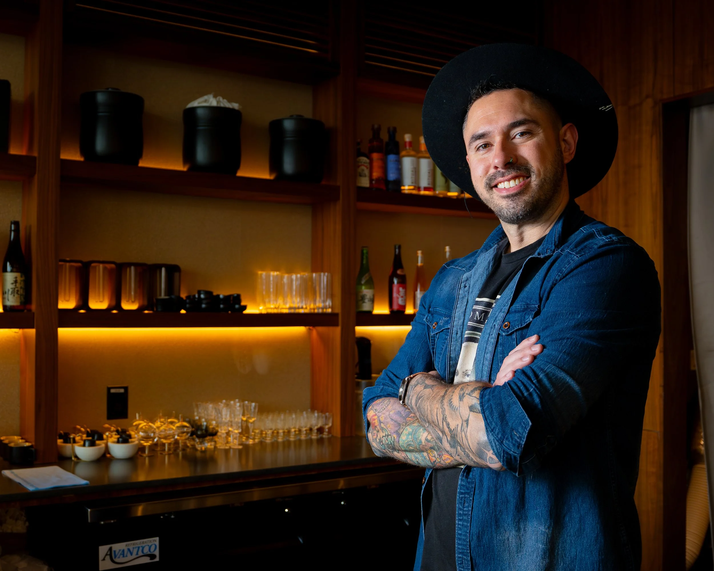 Kosen Tampa chef with tattoos, wearing a black hat and denim jacket, smiling with arms crossed in a bar setting.