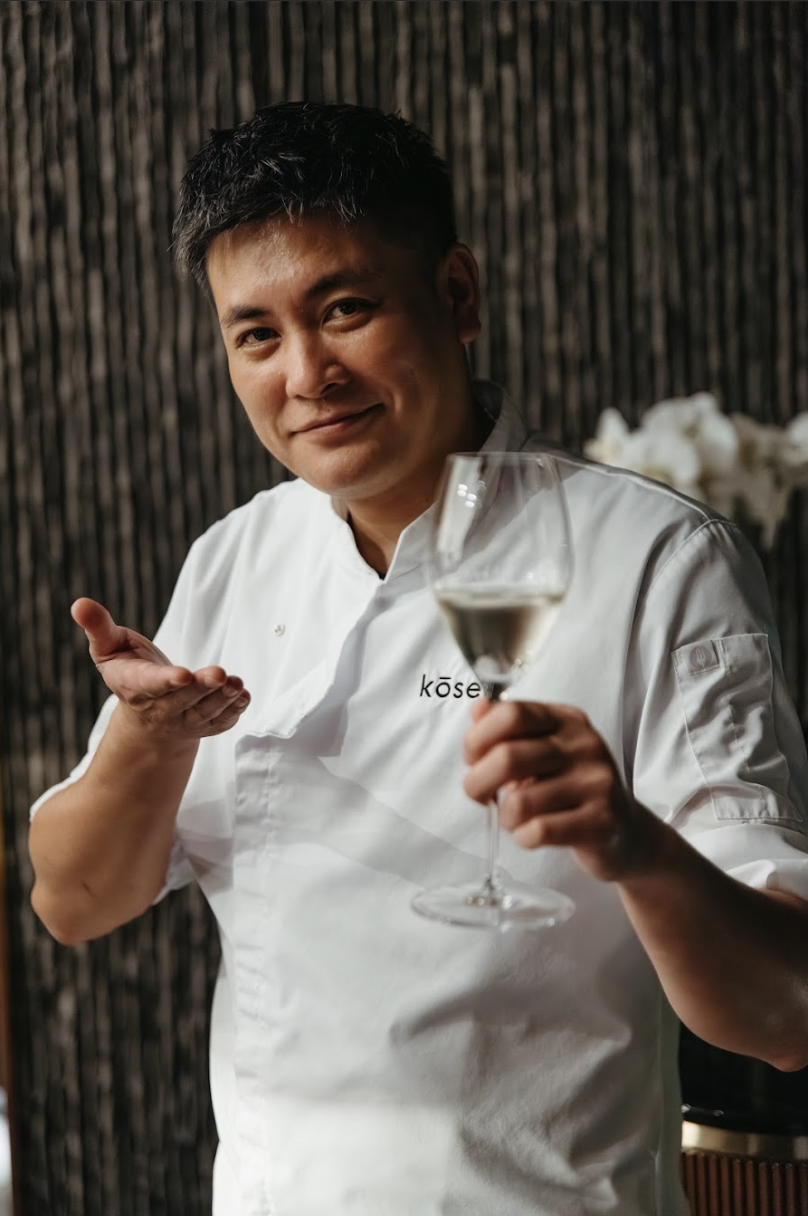Kosen Tampa Chef in a white uniform holding a glass of rosé wine, smiling and gesturing with his hand, in a restaurant setting.