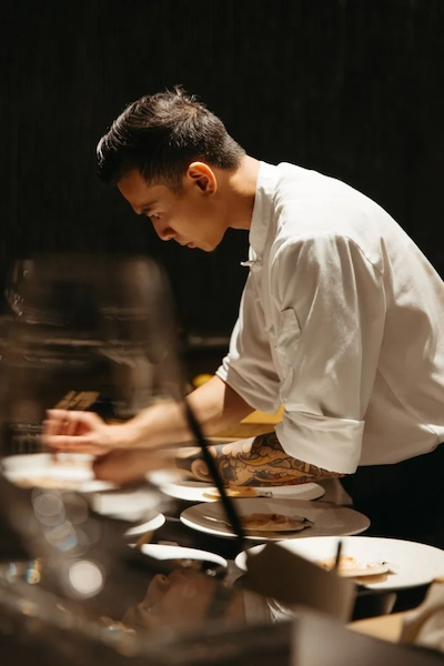 Side profile of a focused chef at Kōsen Tampa, wearing a white coat and featuring traditional Japanese tattoos on his arm, as he meticulously plates a series of dishes under warm, dramatic spotlighting.