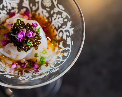 Sliced Mongo Ika (cuttlefish) topped with black caviar and purple shiso flowers, served in an intricate etched glass bowl with a light dashi broth.