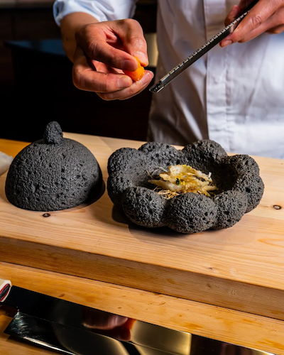 Close-up of a chef’s hands using a microplane to grate fresh citrus or egg yolk over a dish served in a unique, textured black volcanic stone bowl.