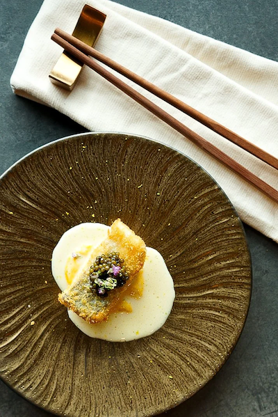 Top-down view of a golden-fried fish fillet topped with a generous portion of black caviar, served on a creamy white sauce on a textured bronze-toned plate.