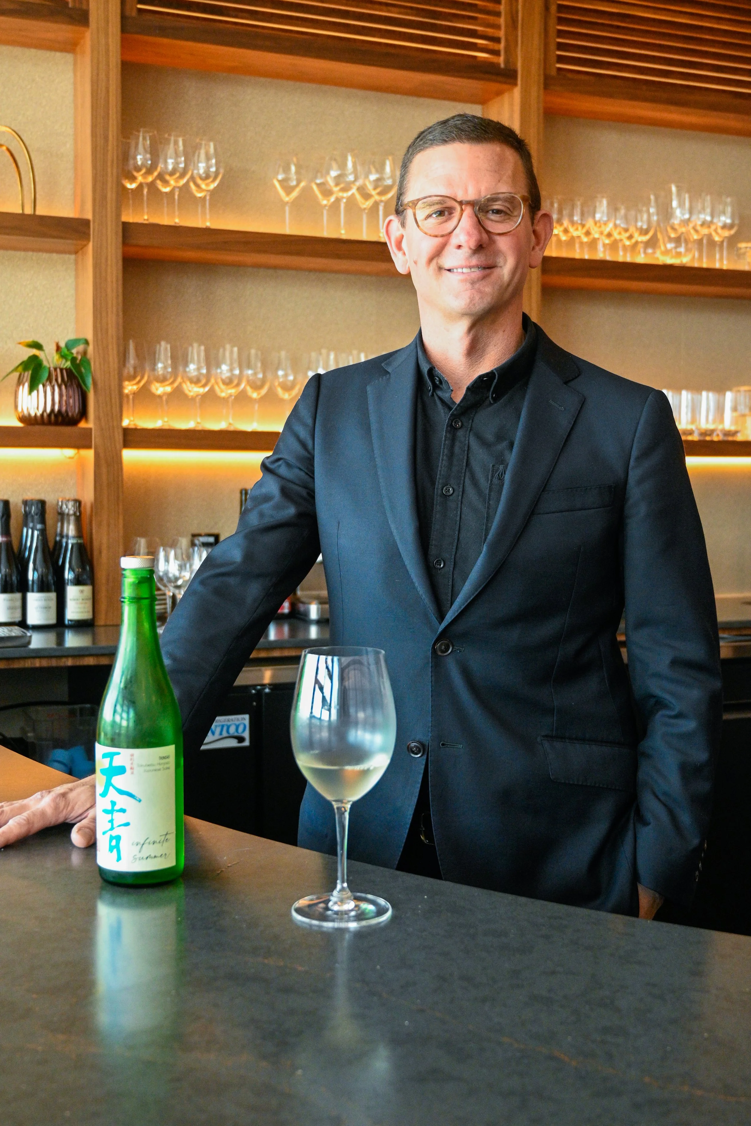 A man in a black suit and glasses smiling at the camera, standing behind a bar with a green sake bottle and a wine glass filled with water on the counter in front of him in Kosen Tampa restaurant.
