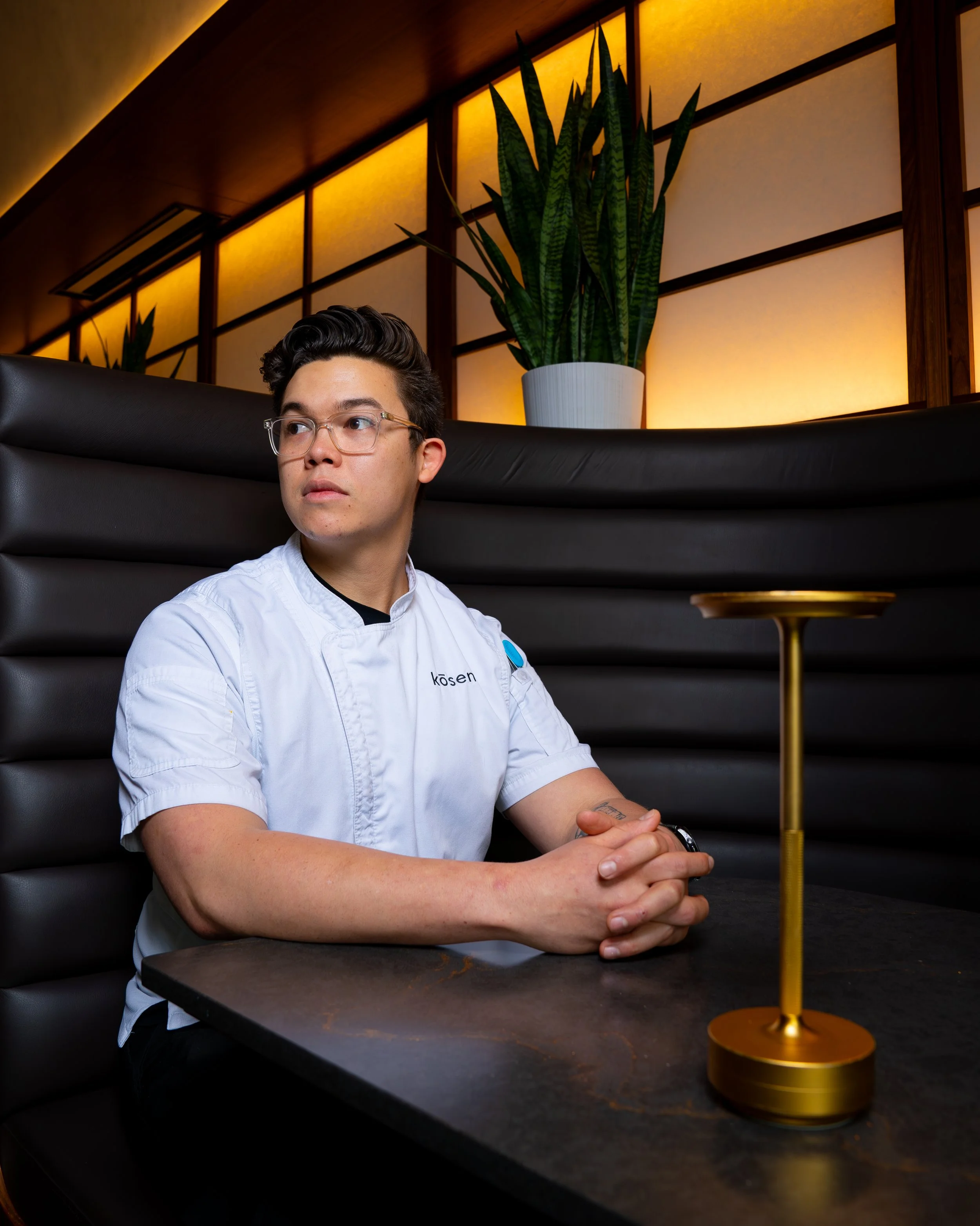 A young man wearing glasses and a white chef's coat with the name tag "kosen" sitting at a dark table in Kosen Tampa. There is a small, modern gold candle holder on the table, and a large potted plant with tall, green leaves behind him. 
