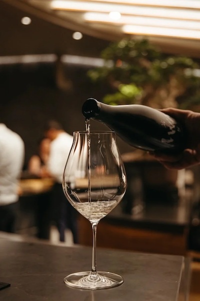 A close-up of clear sake being poured into a tall wine glass on a dark stone counter, with a blurred bonsai tree and warm lighting in the background.