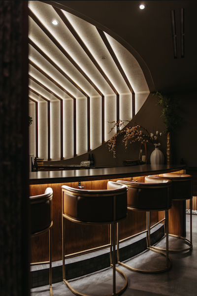 A wide shot of the elegant omakase bar at Kōsen in Tampa, featuring curved leather bar stools with gold frames. The background is dominated by the signature "Ray of Light" architectural wood ribbing and a minimalist floral arrangement.