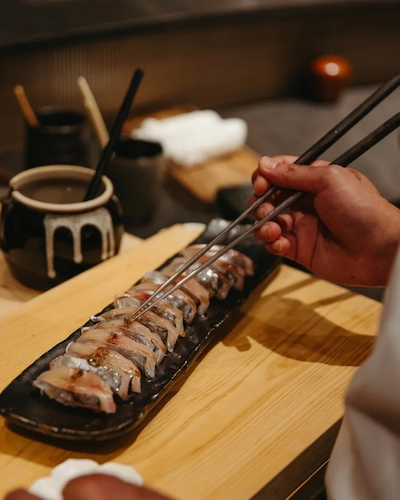 Close-up of a chef’s hands using long metal chopsticks to apply a final garnish to a row of fresh silver-skinned fish nigiri (likely Kohada or Aji) resting on a black ceramic platter over a light wood cutting board.