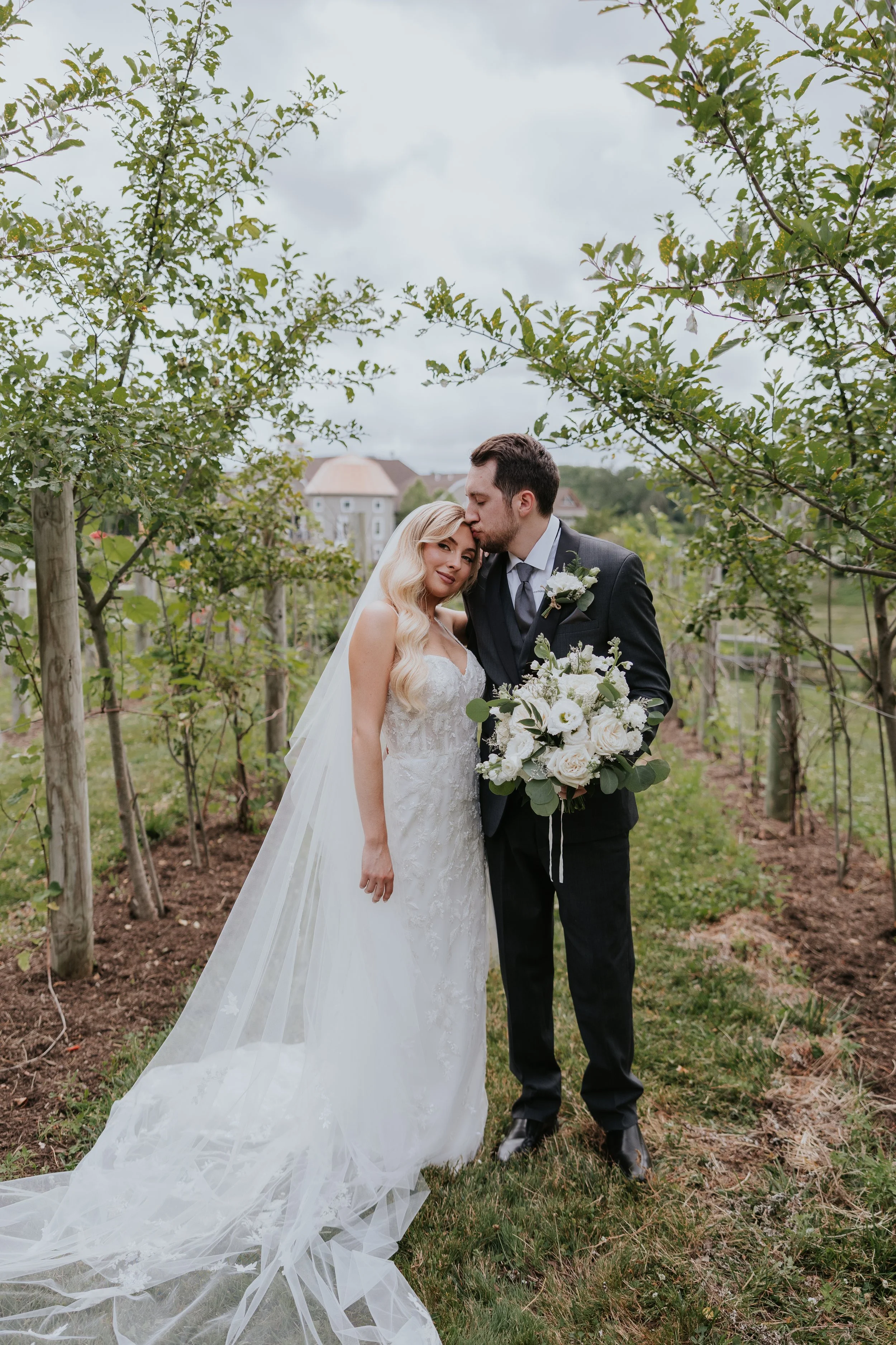 A bride in a white wedding dress with lace details and a groom in a black suit surrounded by green foliage under a cloudy sky.