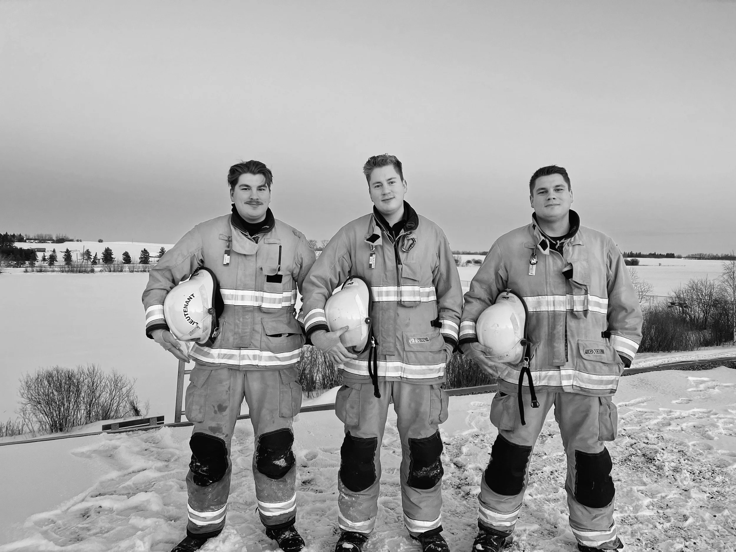 Three firefighters in protective gear standing outdoors in a snowy landscape with trees and fields in the background.