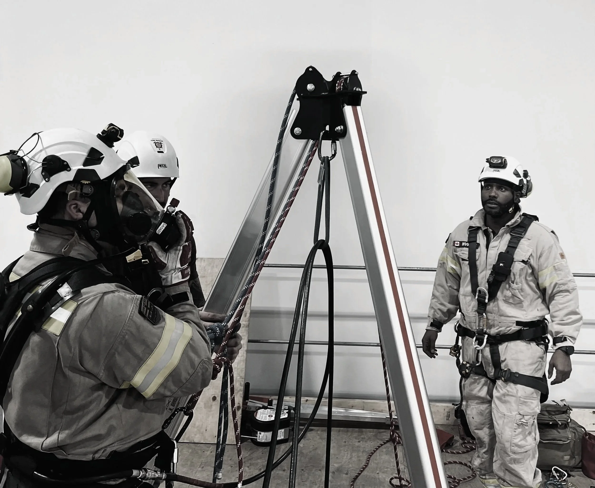 Three rescue workers in gear preparing for a high-angle rescue, with ropes and a pulley system in an indoor setting.