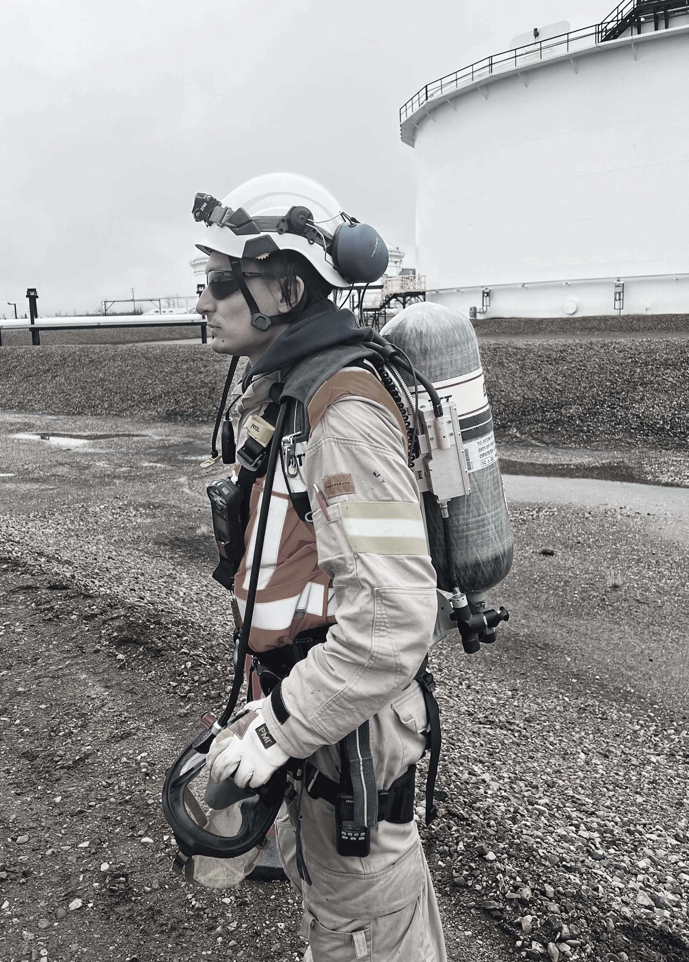 A utility worker wearing protective gear, including a helmet with a headlamp and ear protection, gloves, and a reflective uniform, standing on rocky ground near a large industrial tank or storage facility.