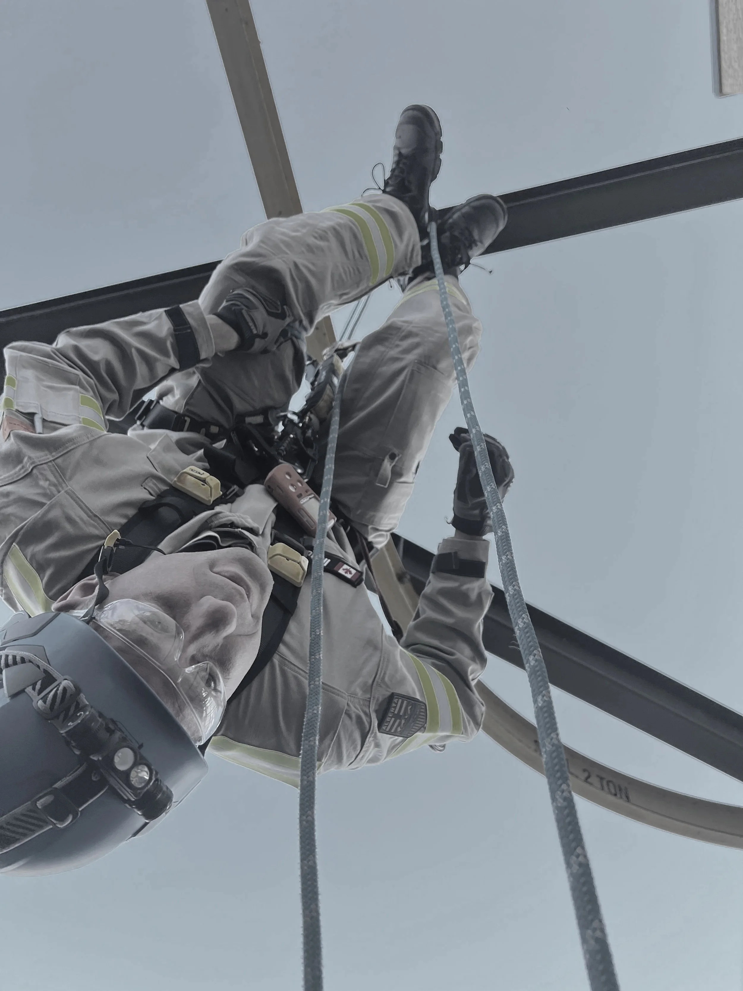 A utility worker in safety gear cleaning high overhead glass windows using a harness and rope.