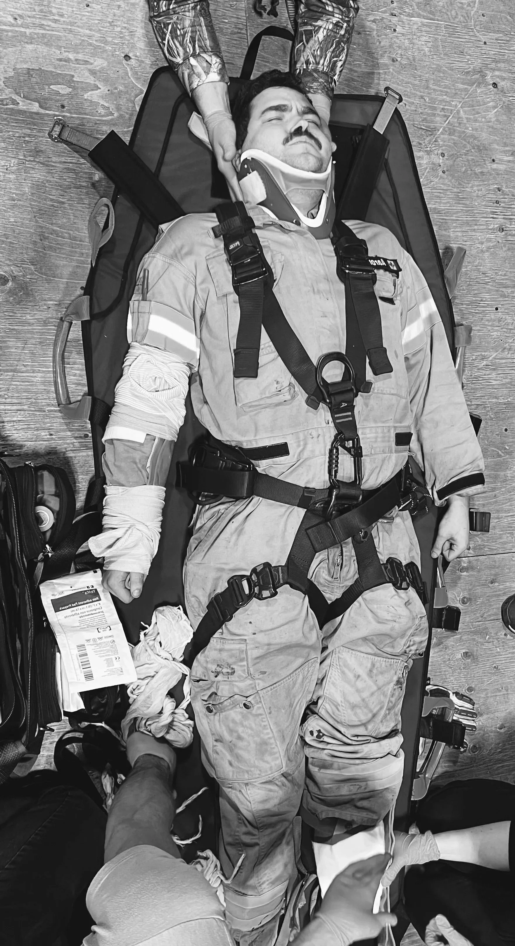 A firefighter lying on a stretcher with his eyes closed, receiving medical assistance. He is dressed in a uniform with a harness, and his arm is wrapped with bandages. Medical supplies and equipment are nearby.