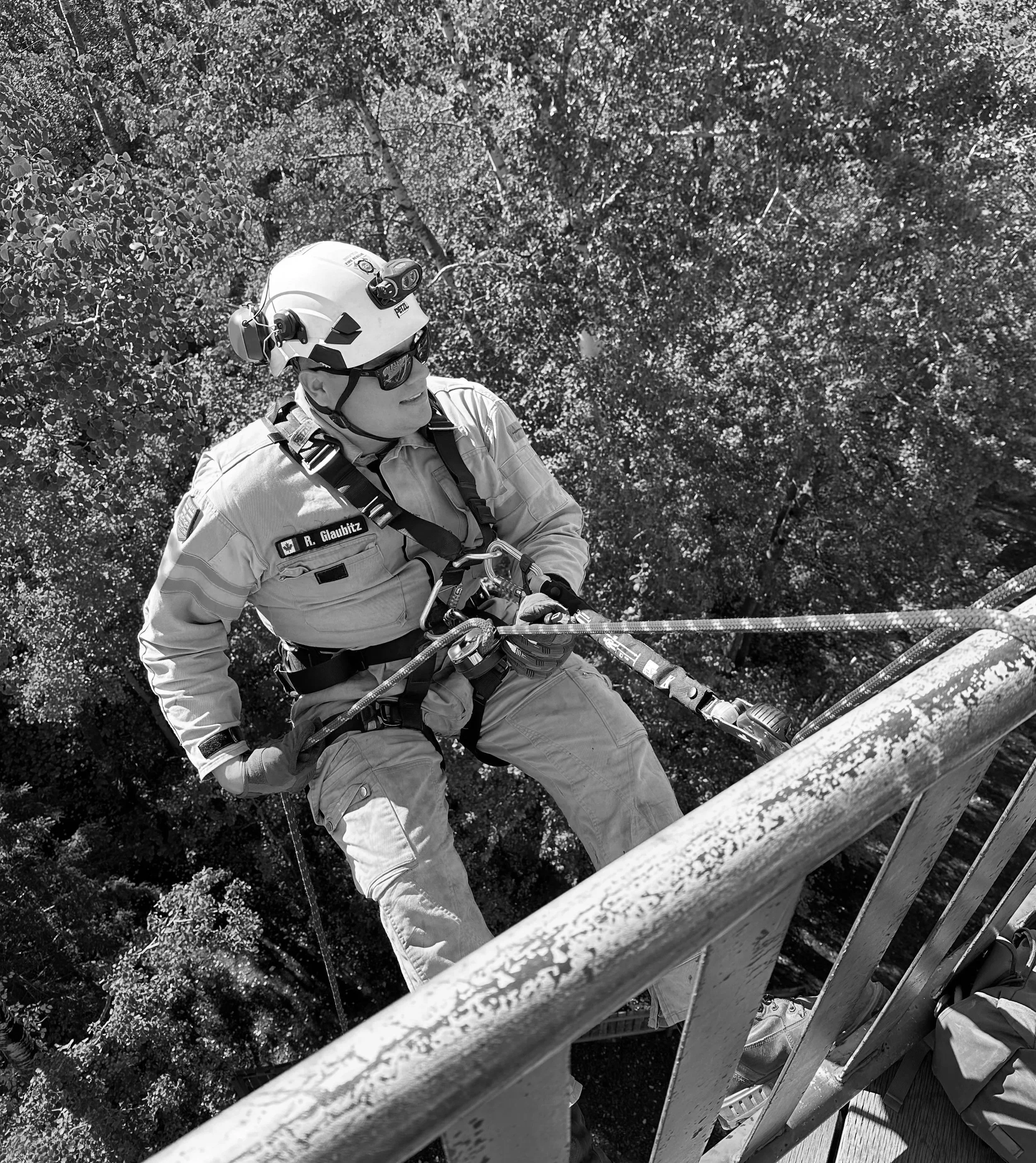 A rescue worker wearing a helmet, safety glasses, and uniform, rappelling down a ladder with trees in the background.