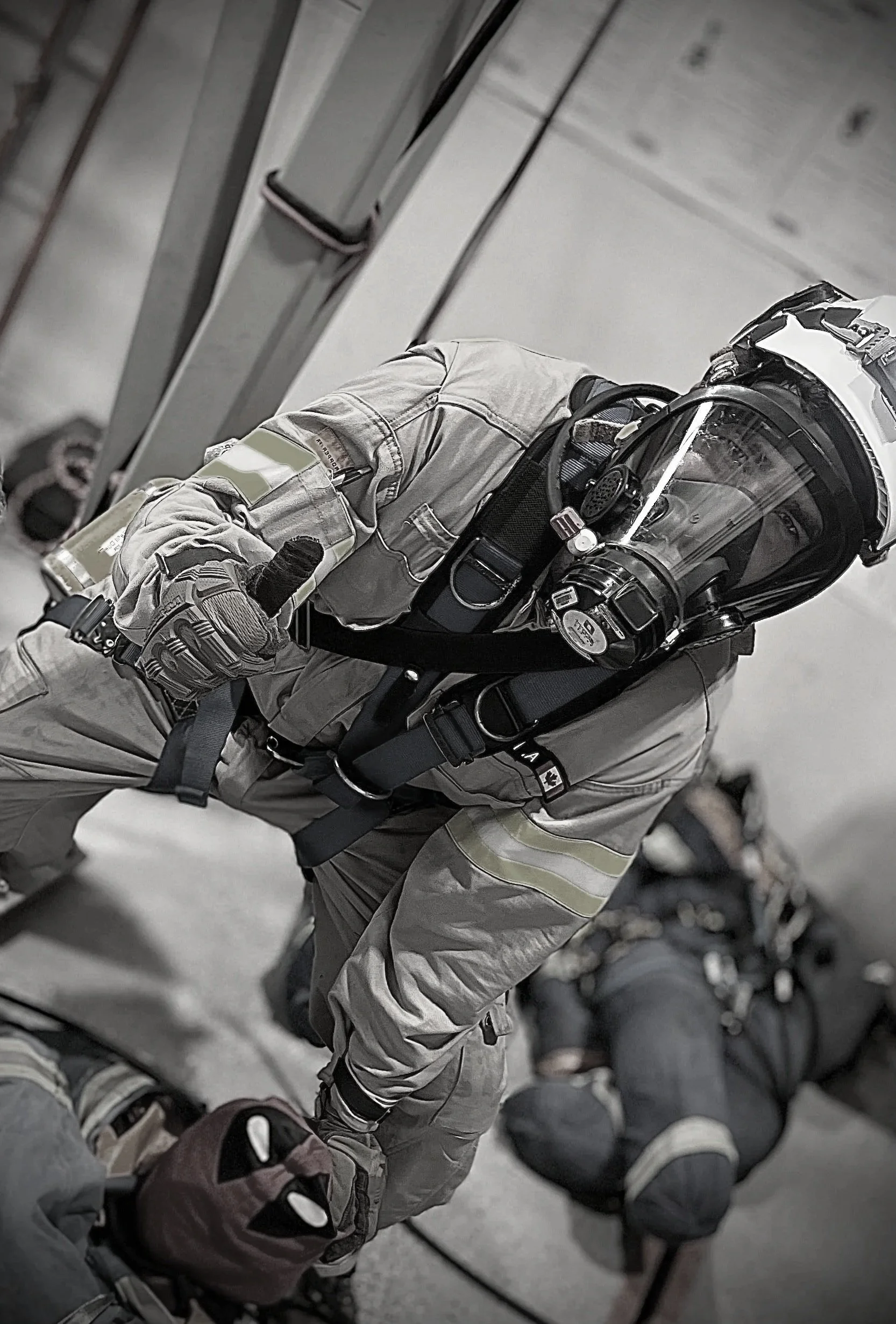 A firefighter in full gear holding a walkie-talkie, kneeling on the ground next to another firefighter, in an indoor setting.