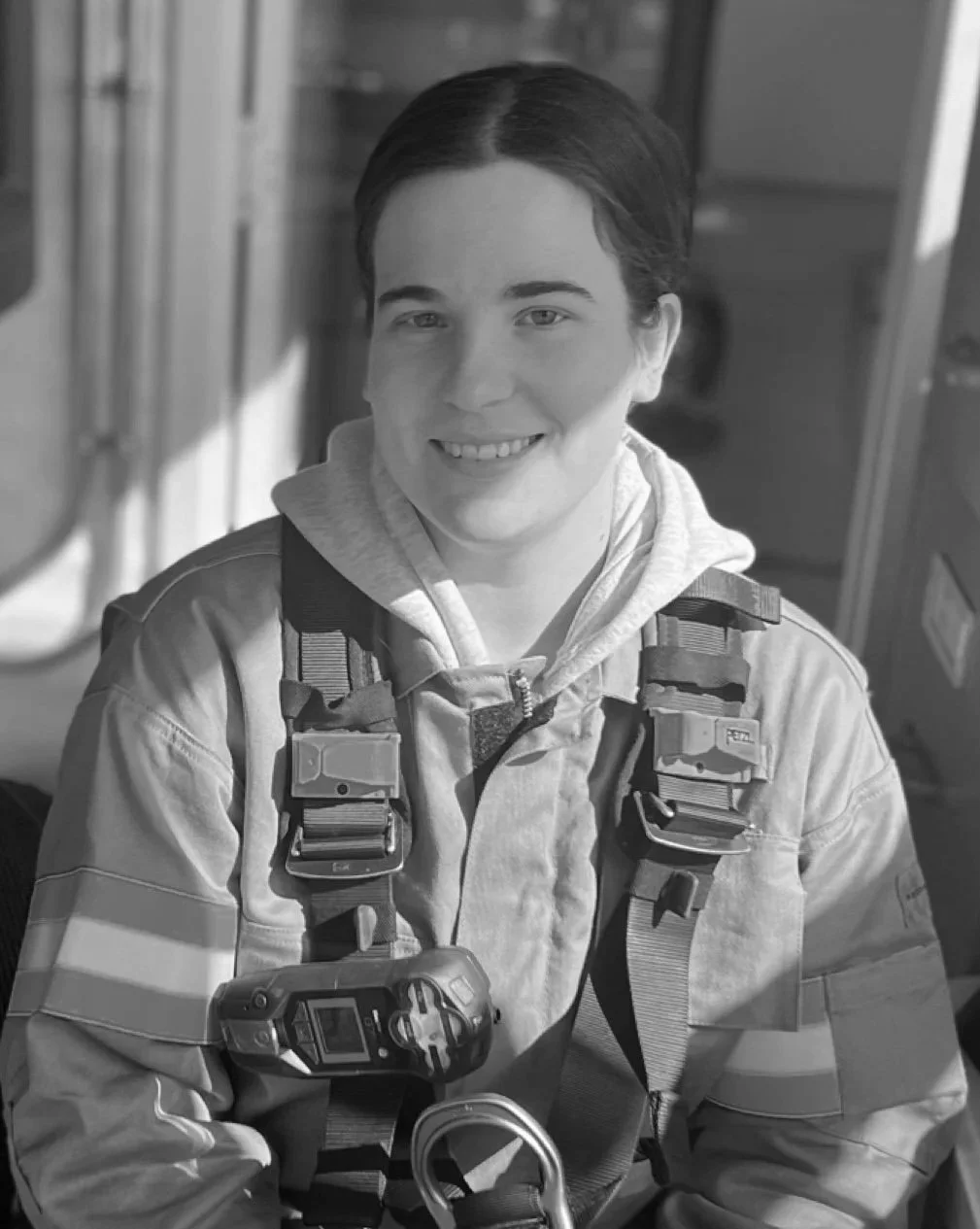 Black and white photo of a young person smiling, wearing a tactical harness and a hoodie, with a camera attached to their gear.