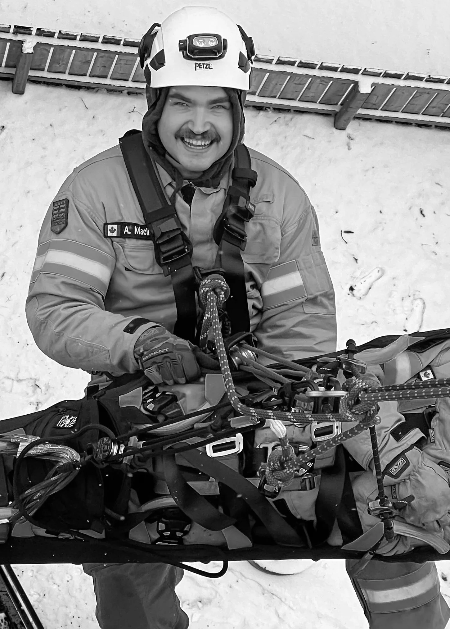 A smiling man wearing a rescue helmet and uniform, standing next to a mountain rescue sled in a snowy environment.