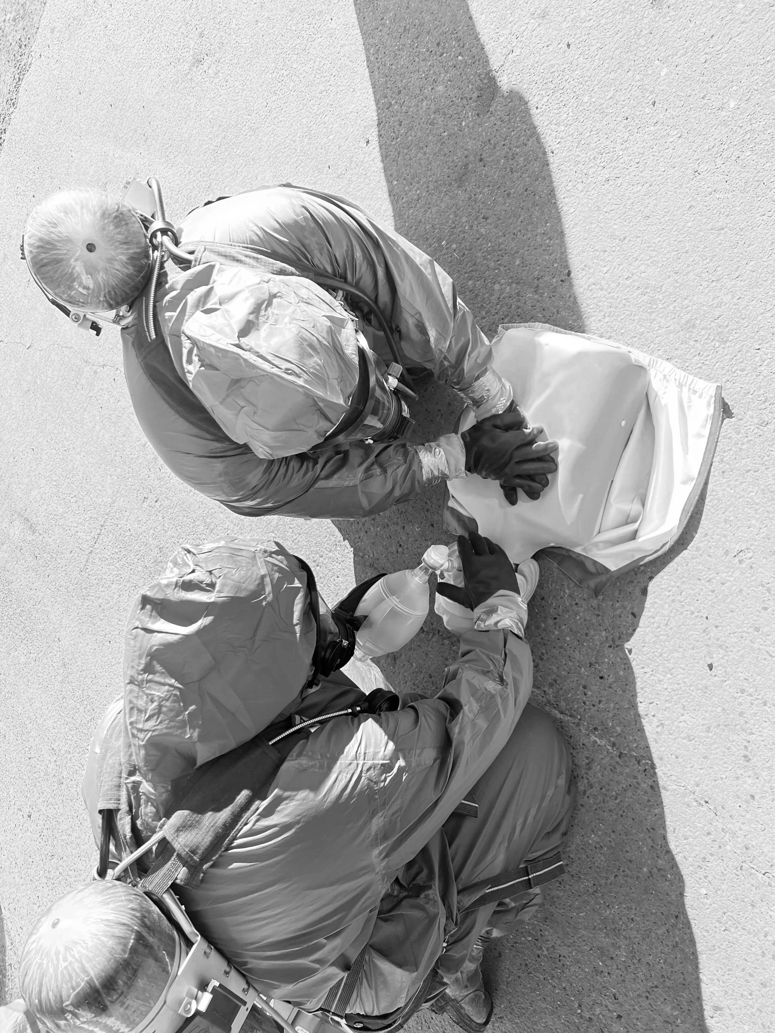 Two individuals in protective hazmat suits kneeling on the ground, handling a large container and a plastic bottle with liquid, possibly engaged in cleanup or scientific work.