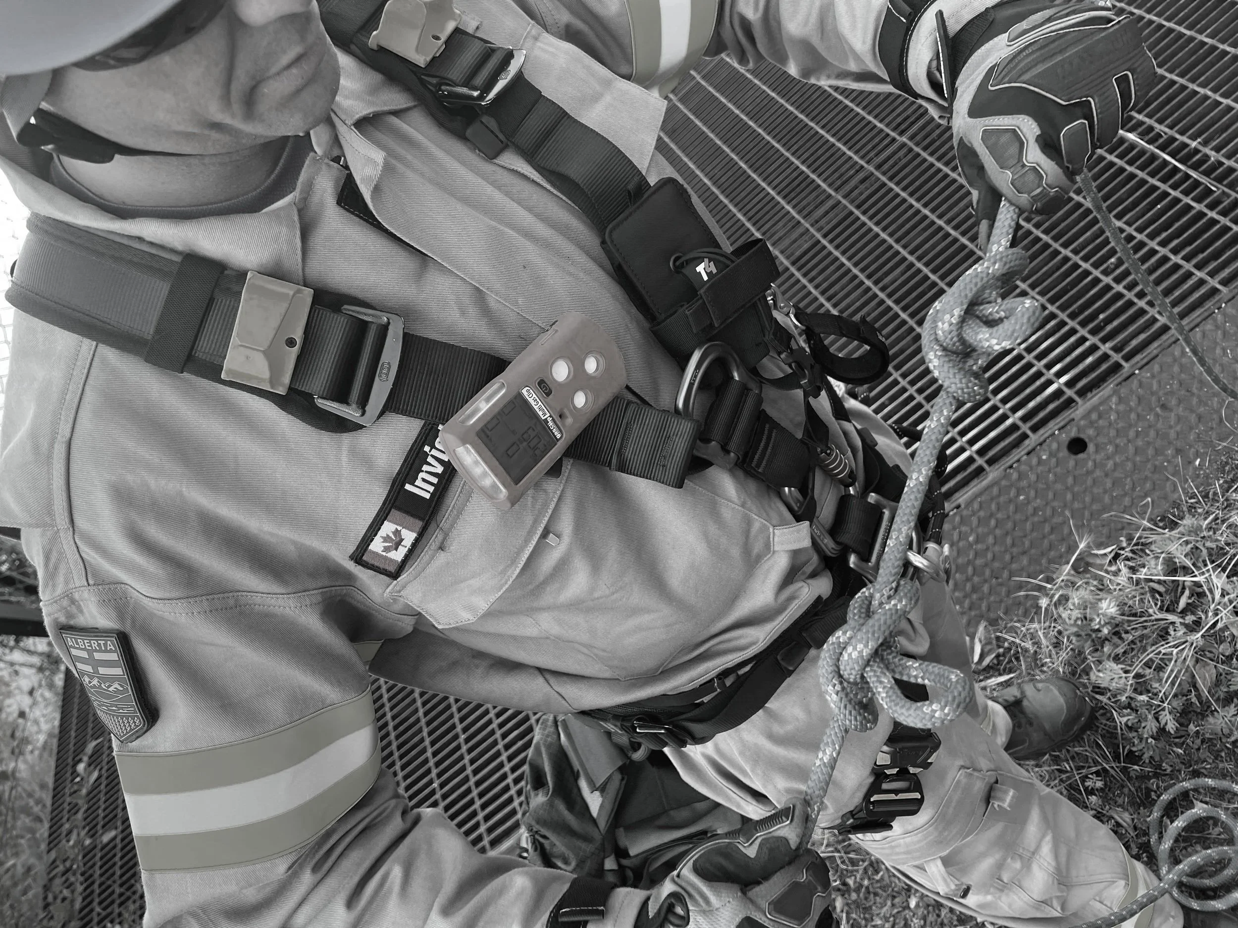 Close-up of a mountain rescue worker wearing gear, including harness, gloves, and climbing equipment, standing on a metal platform with a grating surface and some grass.
