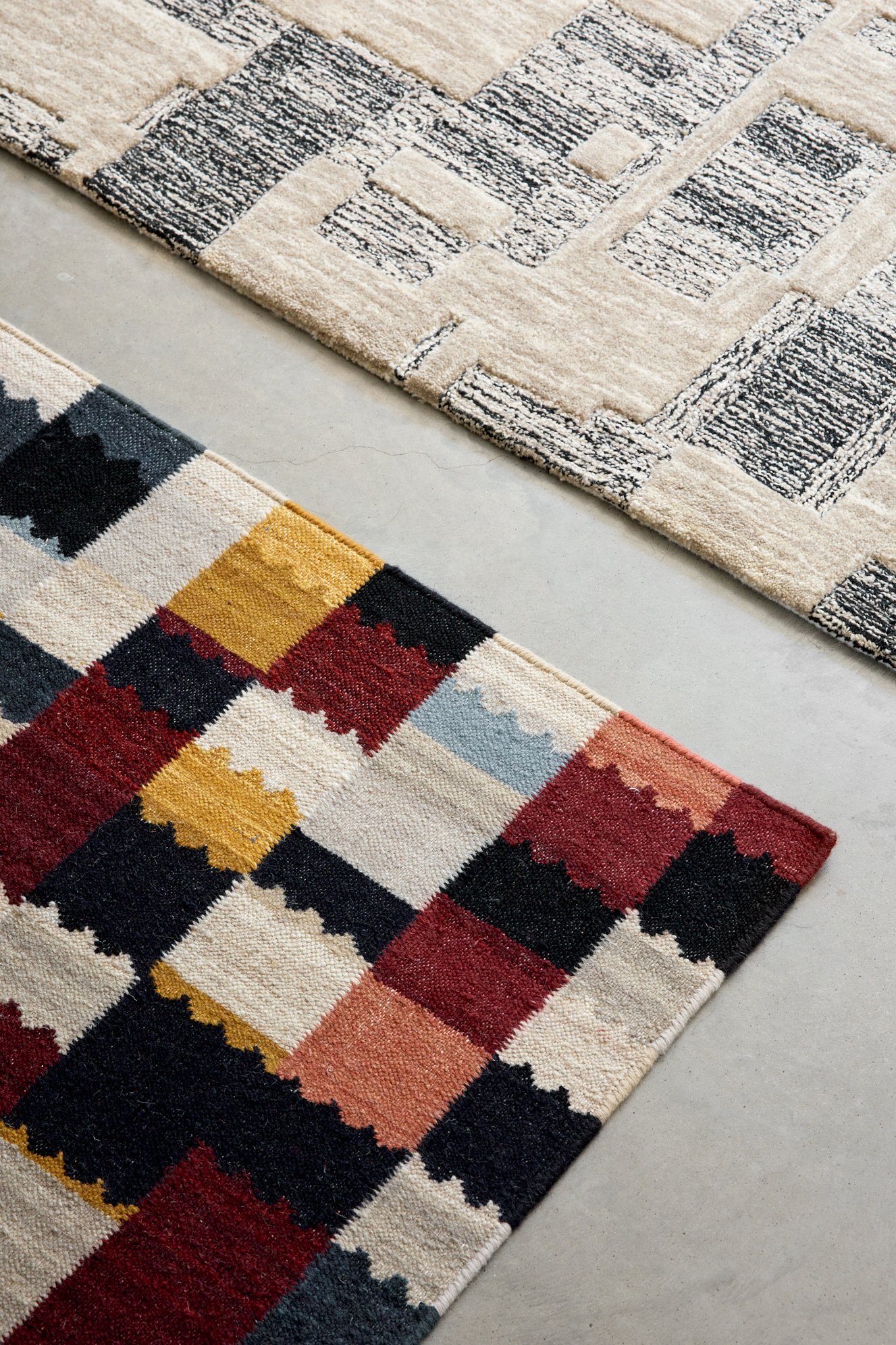 Two rectangular area rugs, one with a patterned design in black, beige, and gray, and the other with a colorful plaid pattern in red, yellow, black, and beige, laid out on a concrete floor.