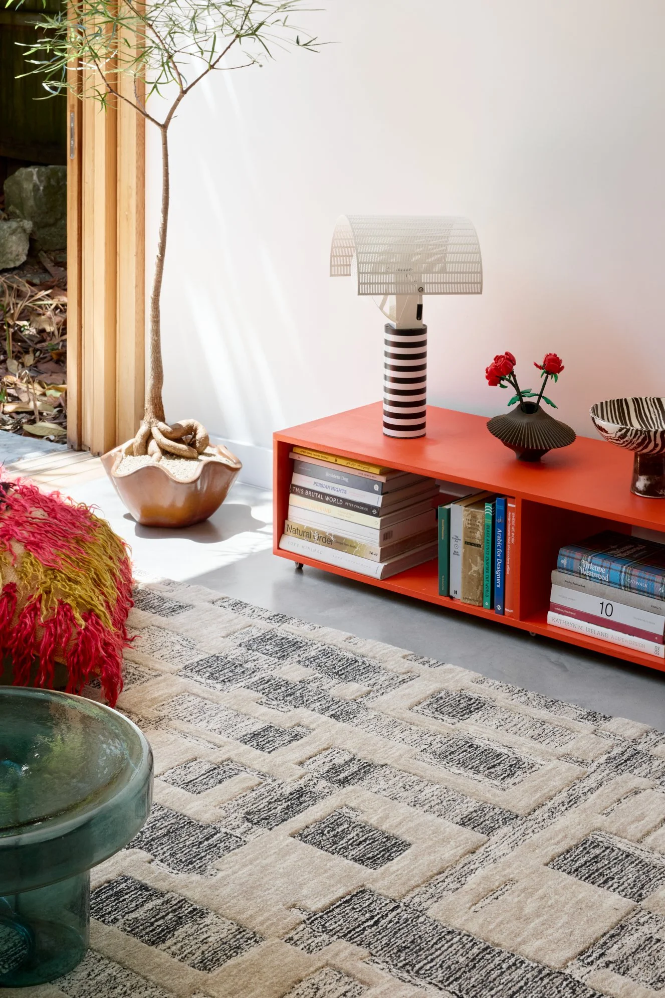 A cozy living room corner with a red bookshelf, a table lamp with a striped base, and a potted plant with twisted trunk near a sliding door with a view of outdoor rocks and leaves. There's a patterned rug and a fuzzy red and yellow ottoman.