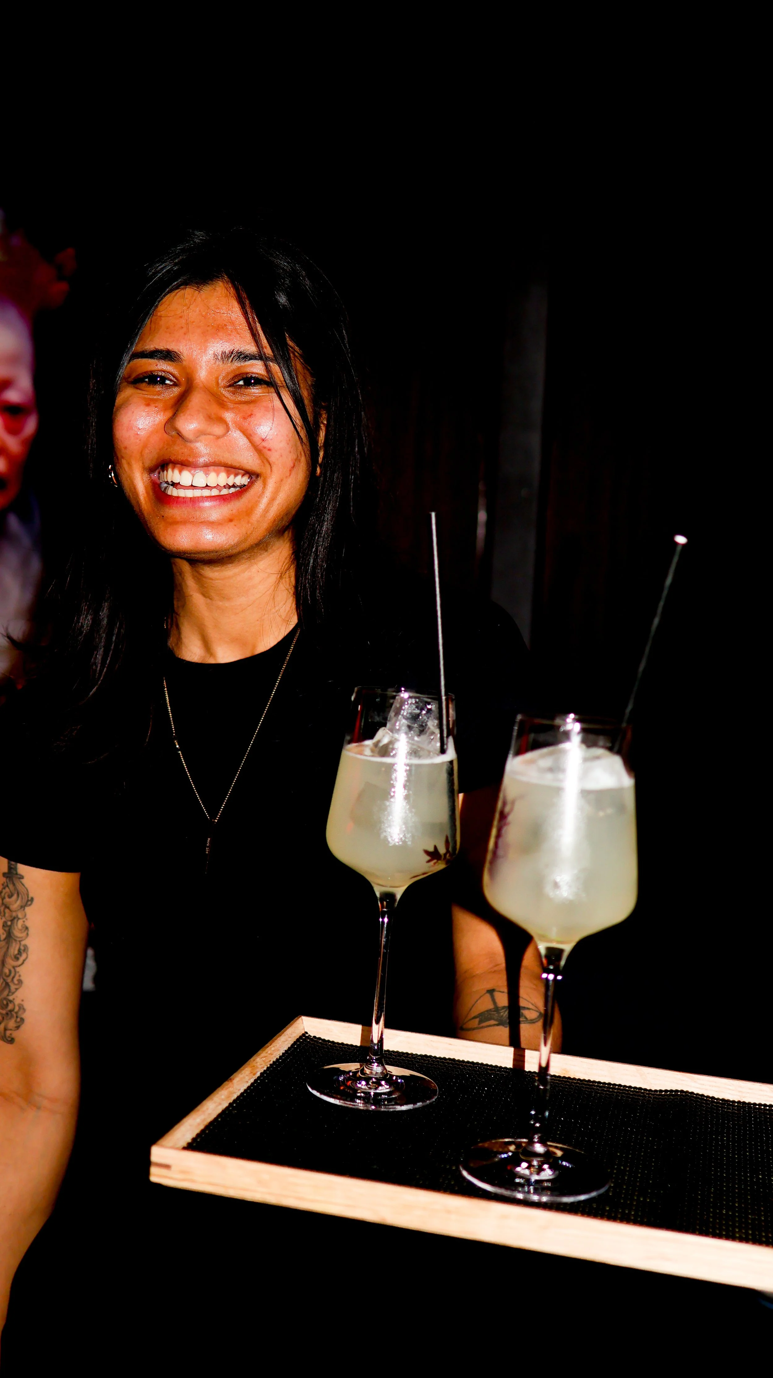 A woman with long black hair smiling, sitting at a bar with two glasses of cocktails on a tray in front of her.