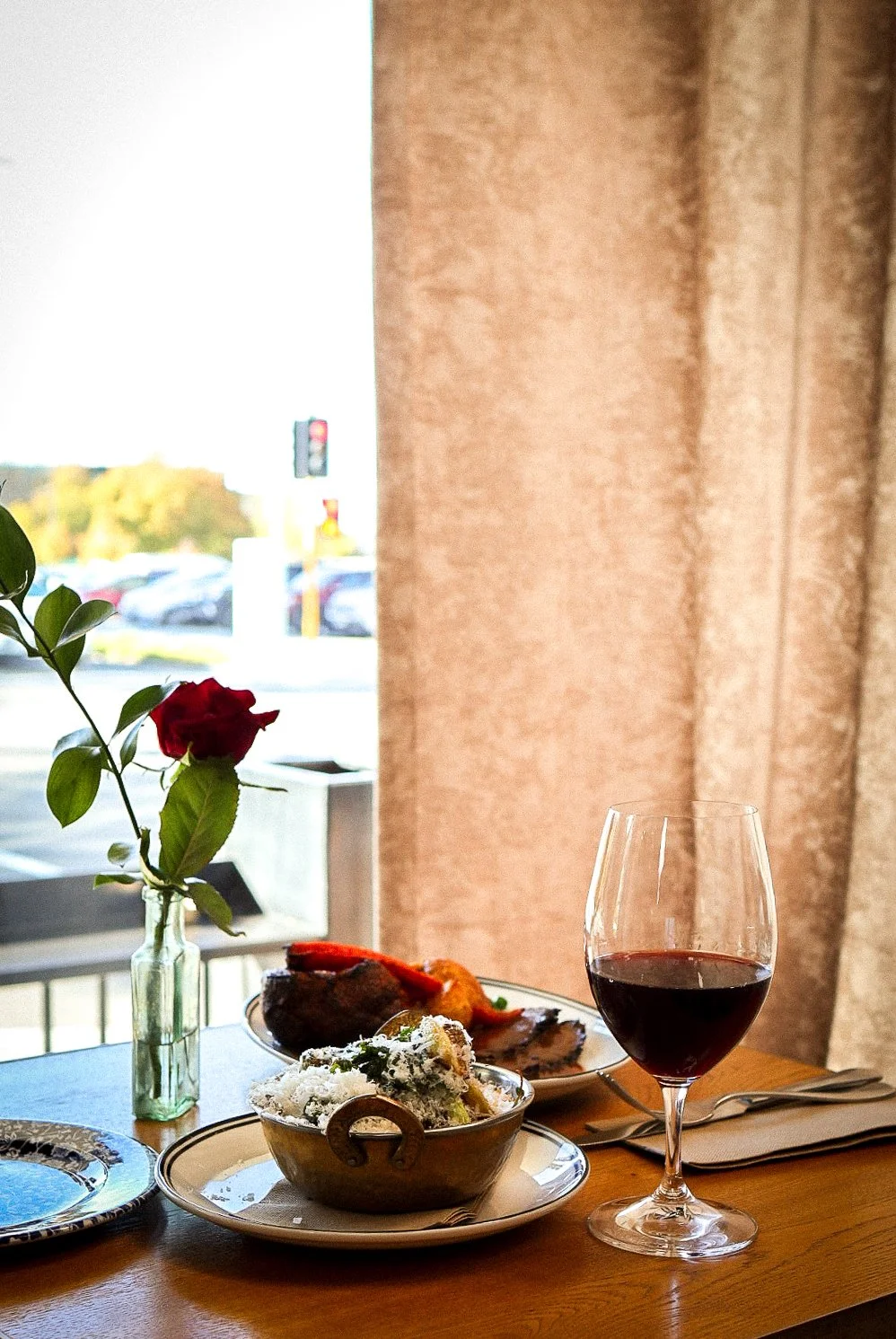 A table with a glass of red wine, a small bowl of food, and a plate with roasted meat and vegetables, next to a vase with a single red rose, near a window with beige curtains.