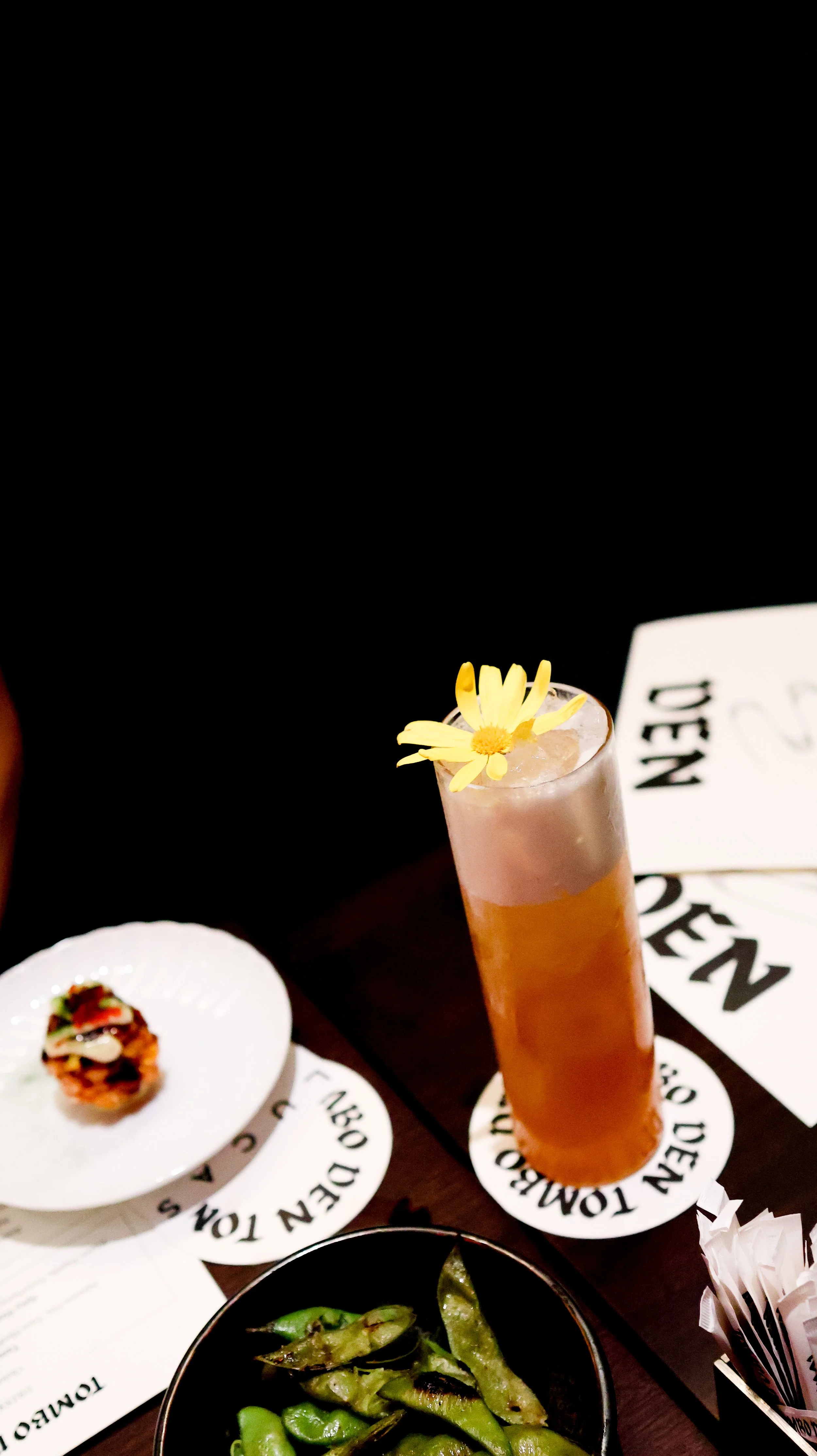 A tall glass drink garnished with a yellow flower, a small plate with a topped appetizer, a bowl of grilled green peppers, and paper coasters with text on a dark table.