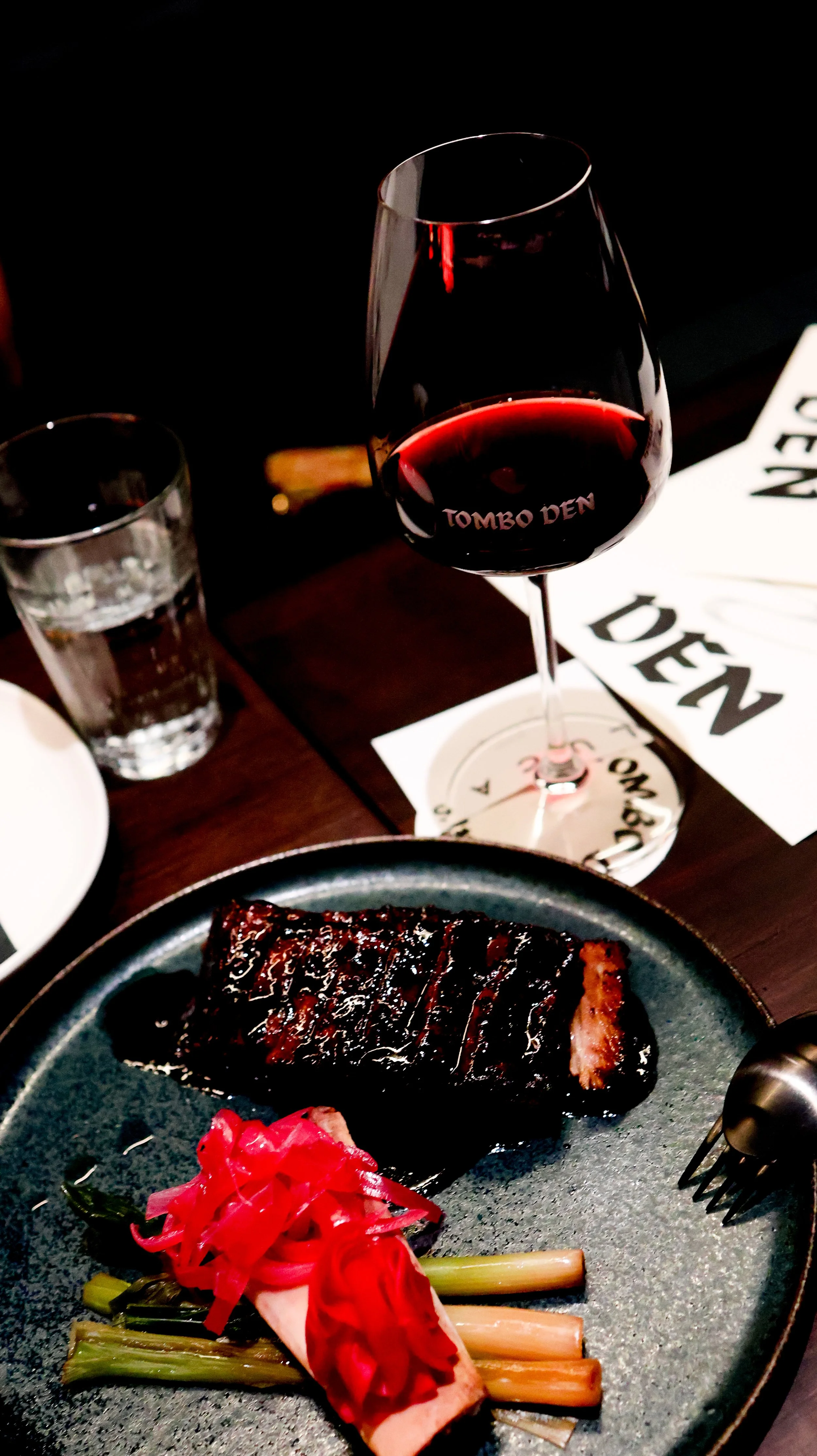 A plate with a glazed, cooked piece of beef, pink pickled vegetables, and green onions, with a glass of red wine, a glass of water, and restaurant napkins on a dark table.