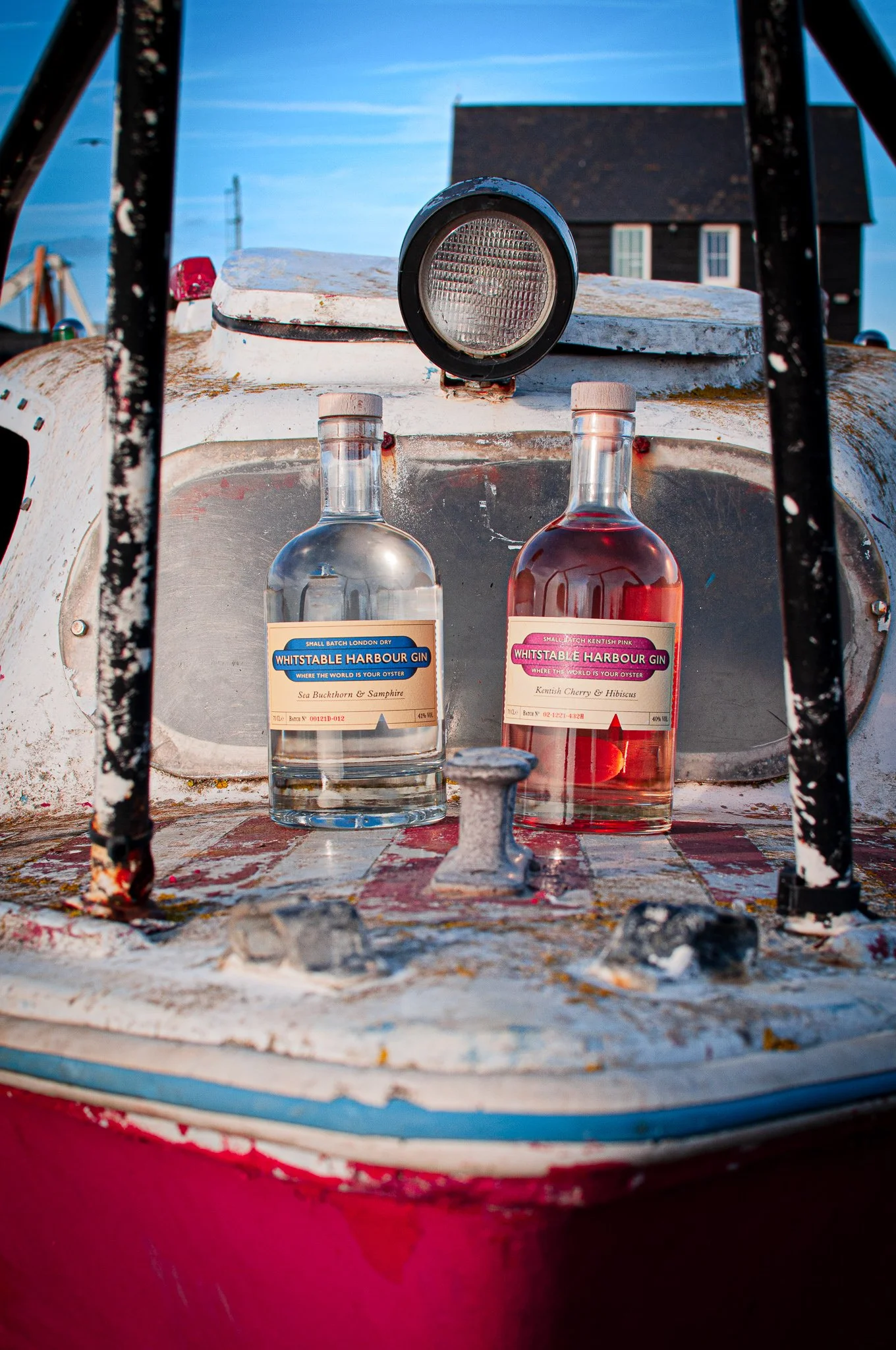 Two bottles of Whitstable Harbour Gin, placed on the deck of an old, weathered boat with a small portlight and a spotlight above, against a background of a blue sky as part of social media and website content that I shot and styled.